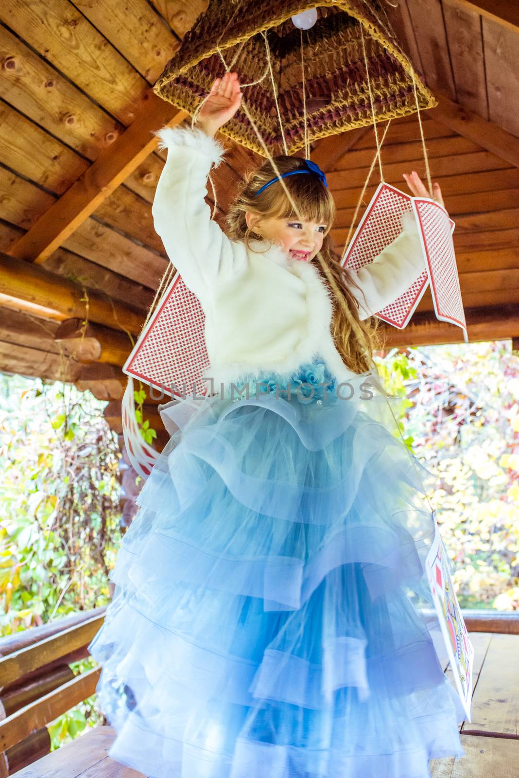 An little beautiful girl in a long blue dress in the scenery of Alice in Wonderland playing and dancing with large playing cards on the table in the garden.
