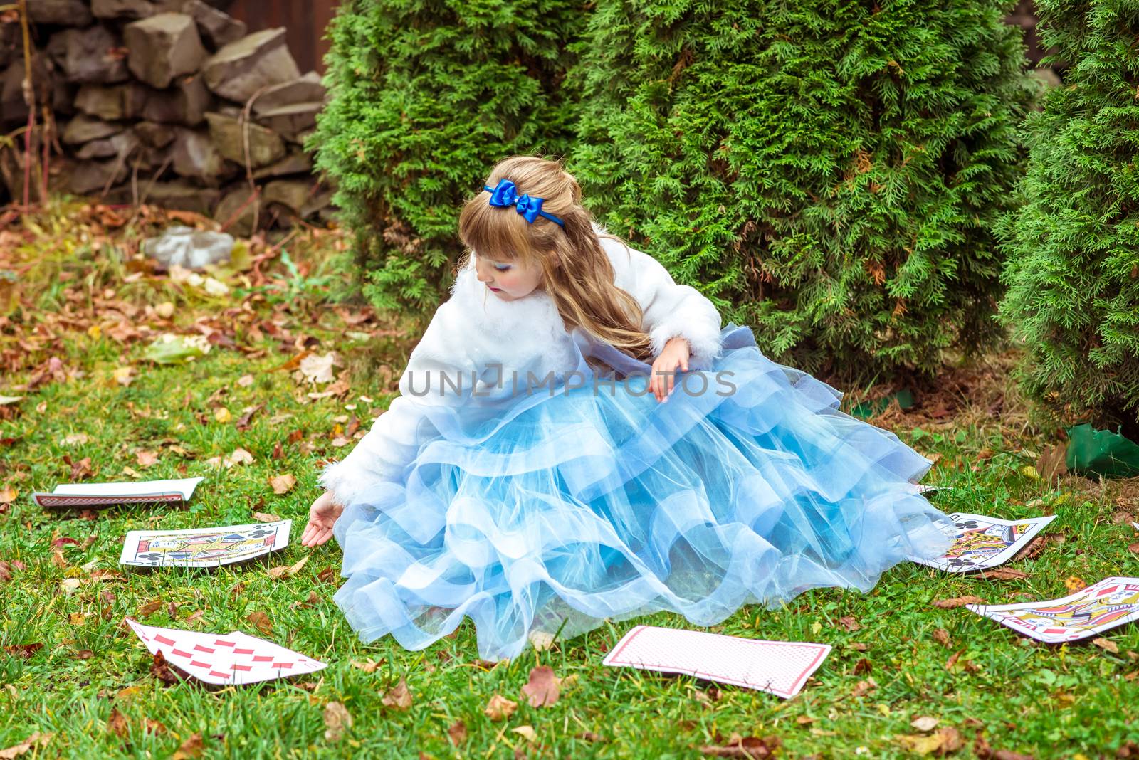 An little beautiful girl in a long blue dress in the scenery of Alice in Wonderland sitting on the grass and playing with large game cards.