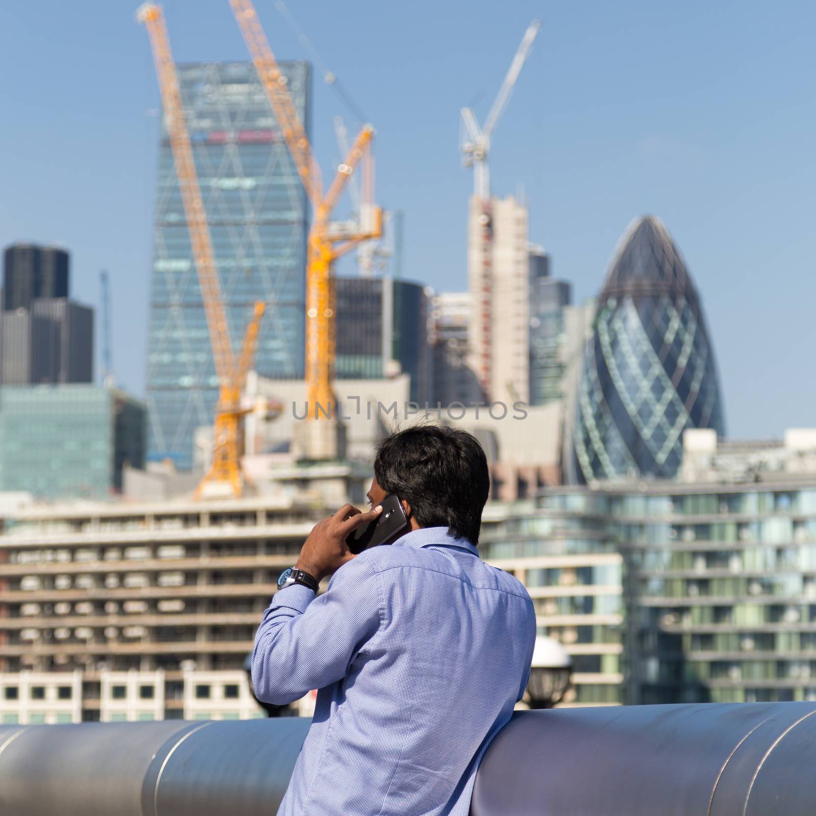 International businessman talking on mobile phone outdoor, looking at modern skyscrapers in London city, UK.