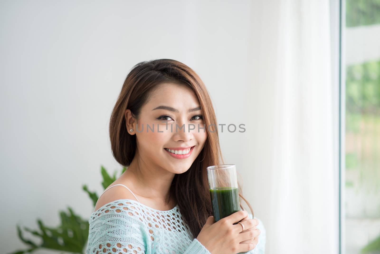 Smiling young asian woman drinking green fresh vegetable juice or smoothie from glass at home