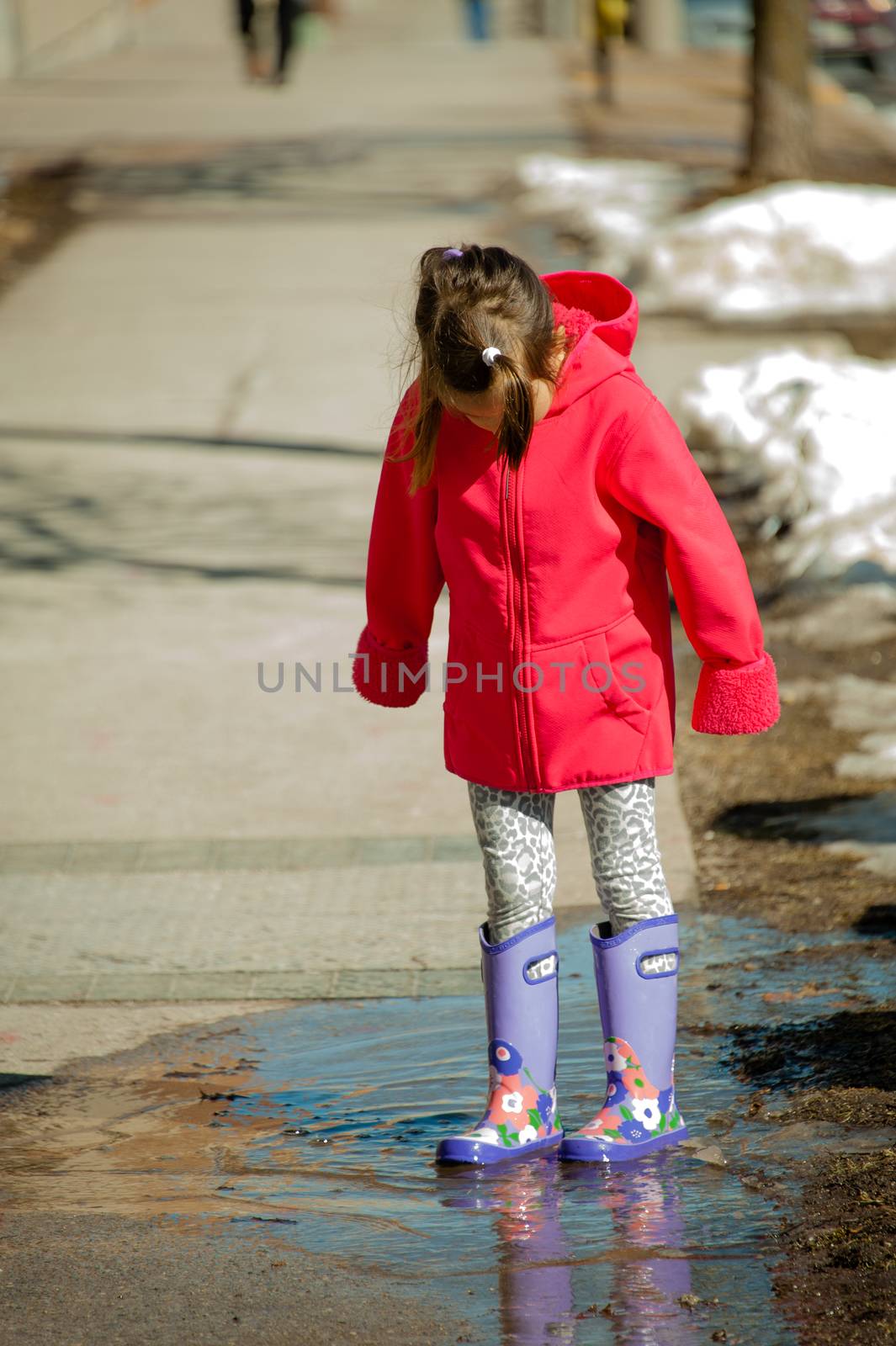 A girl in a red jacket and rubber boots is standing in a spring puddle of melted snow
