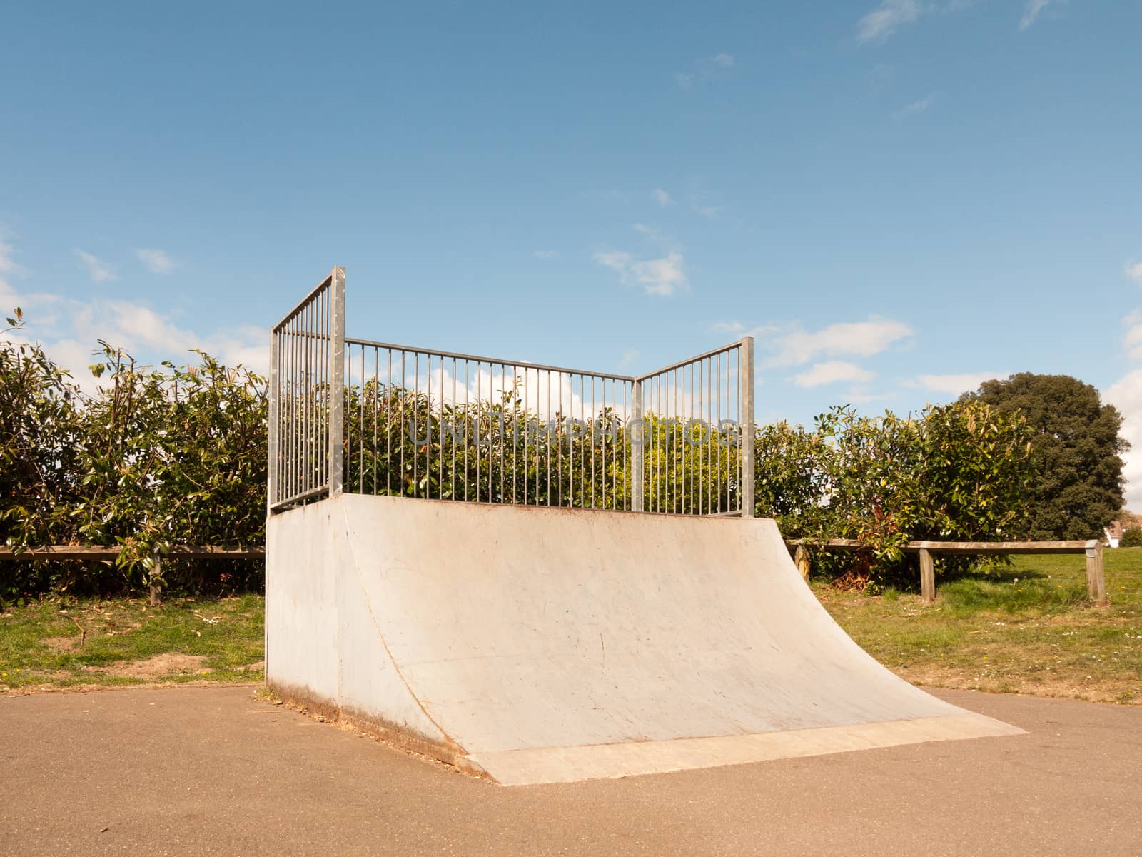 An Empty and Unused Ramp Half Pipe at the Skate Park in the Country Park in the UK Shining in the Sunlight of the Day and with A Shrub Hedge Behind the Bars Protecting it Fenced in the Spring in the UK