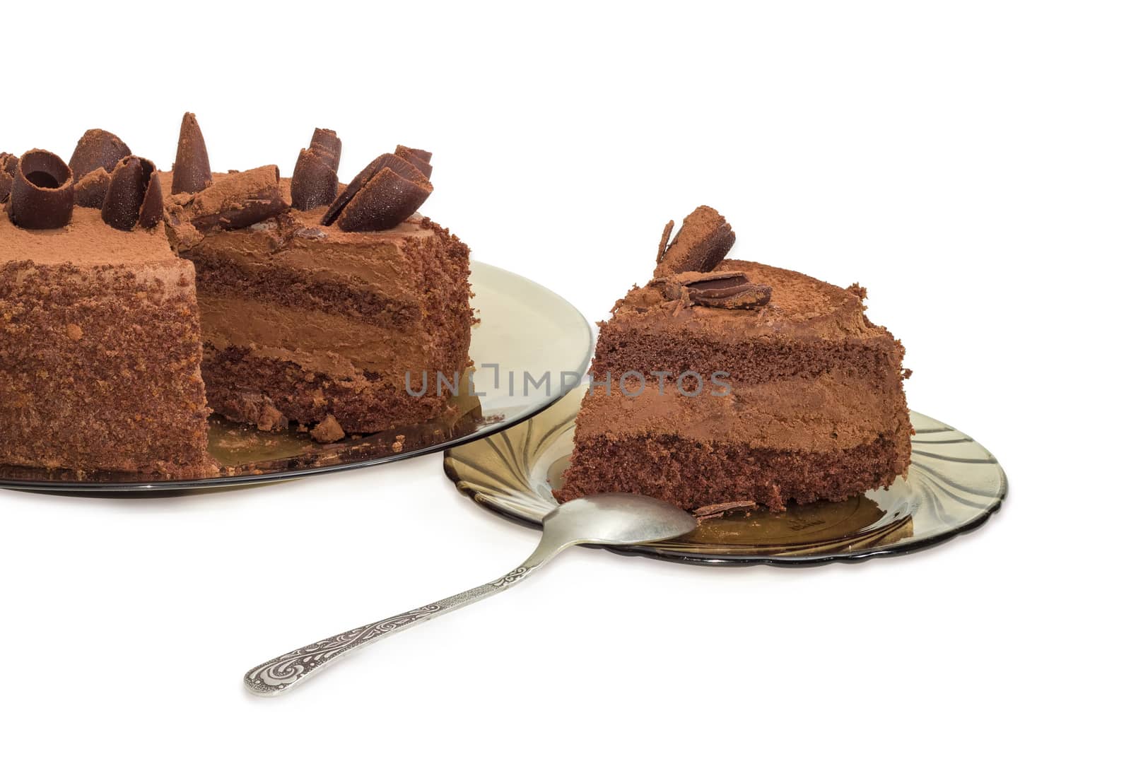 Partly sliced round layered chocolate cake decorated with chocolate chips and sprinkled with cocoa powder on the dark glass dish and slice cake on saucer with spoon on a light background
