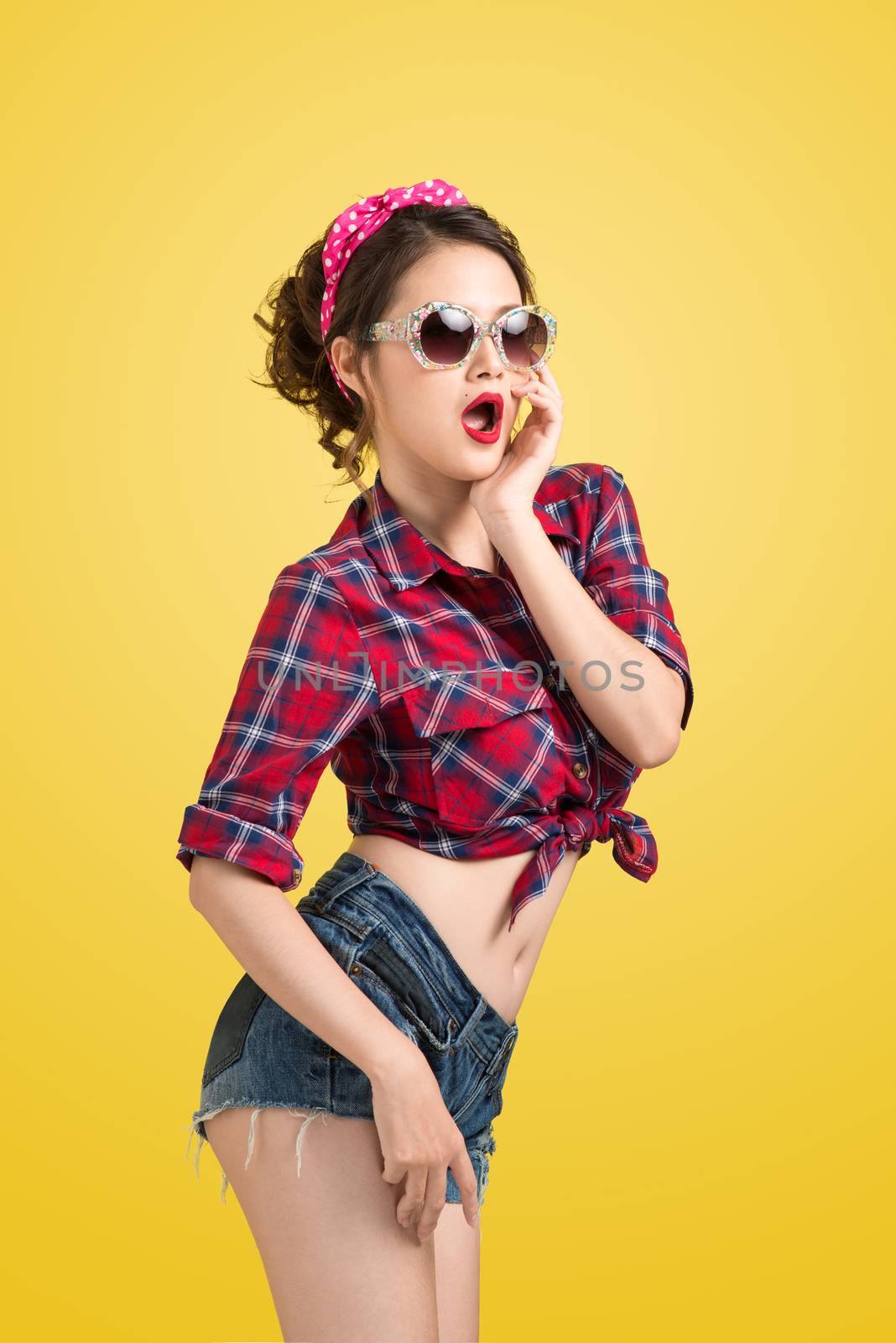 Lovely woman retro portrait with pin-up make-up and hairstyle wearing sunglasses posing over yellow background.