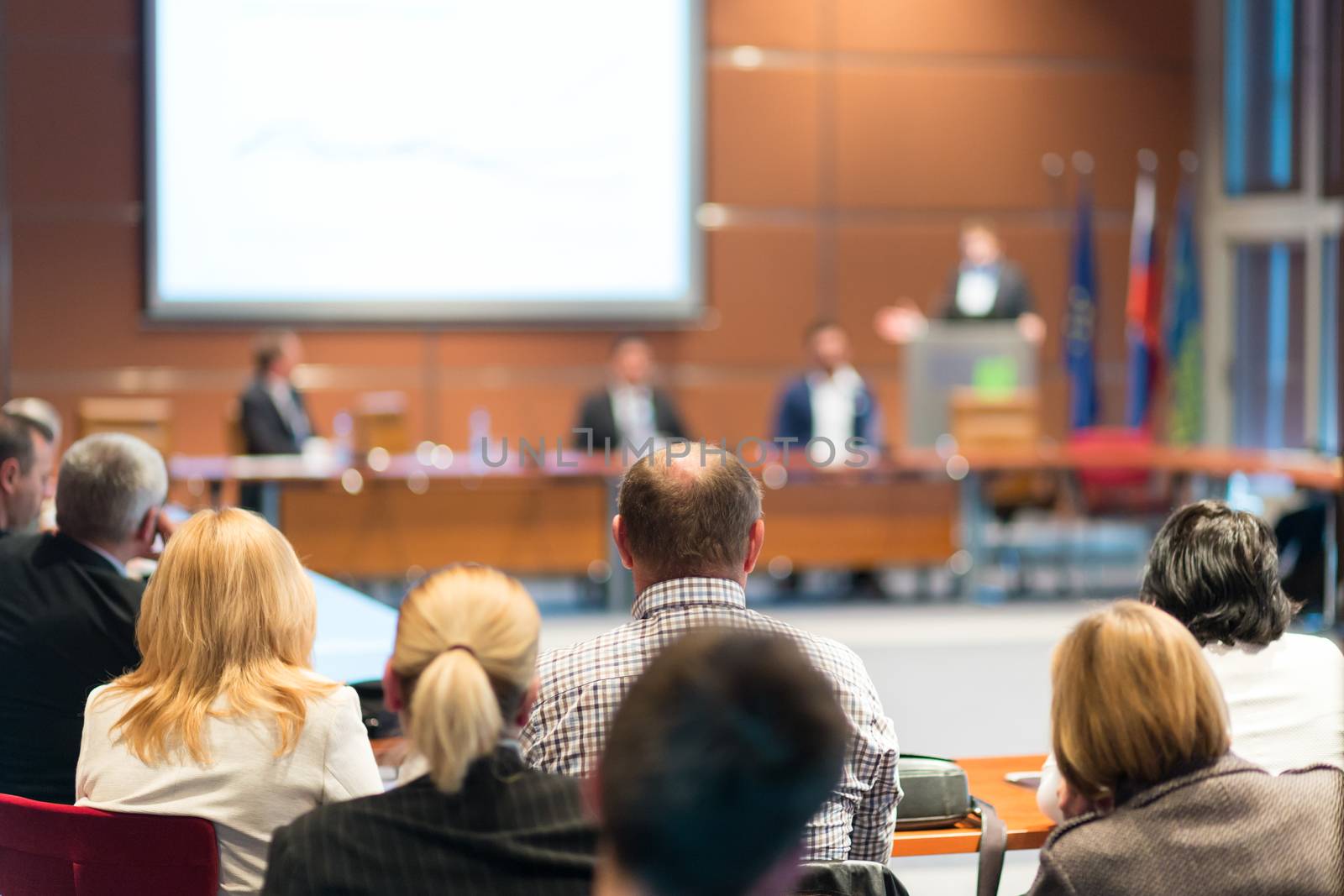 Speaker giving a talk on corporate business event. Audience at the conference hall. Business and Entrepreneurship event. Focus on unrecognizable business people in the rear.