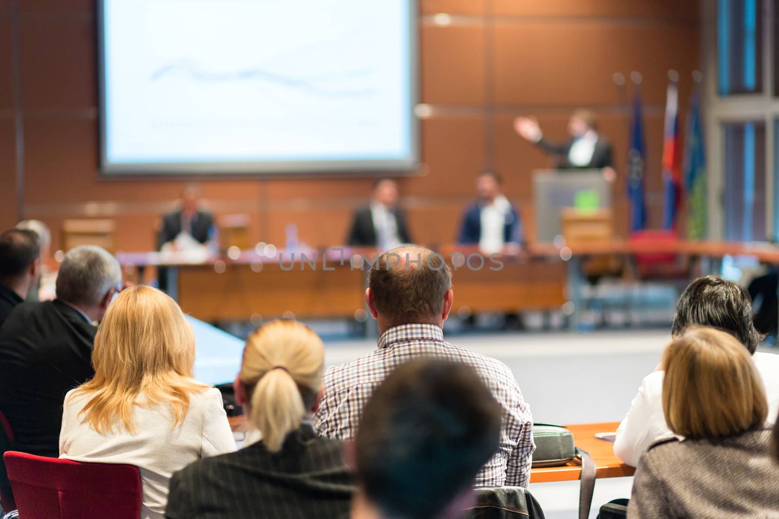 Speaker giving a talk on corporate business event. Audience at the conference hall. Business and Entrepreneurship event. Focus on unrecognizable business people in the rear.