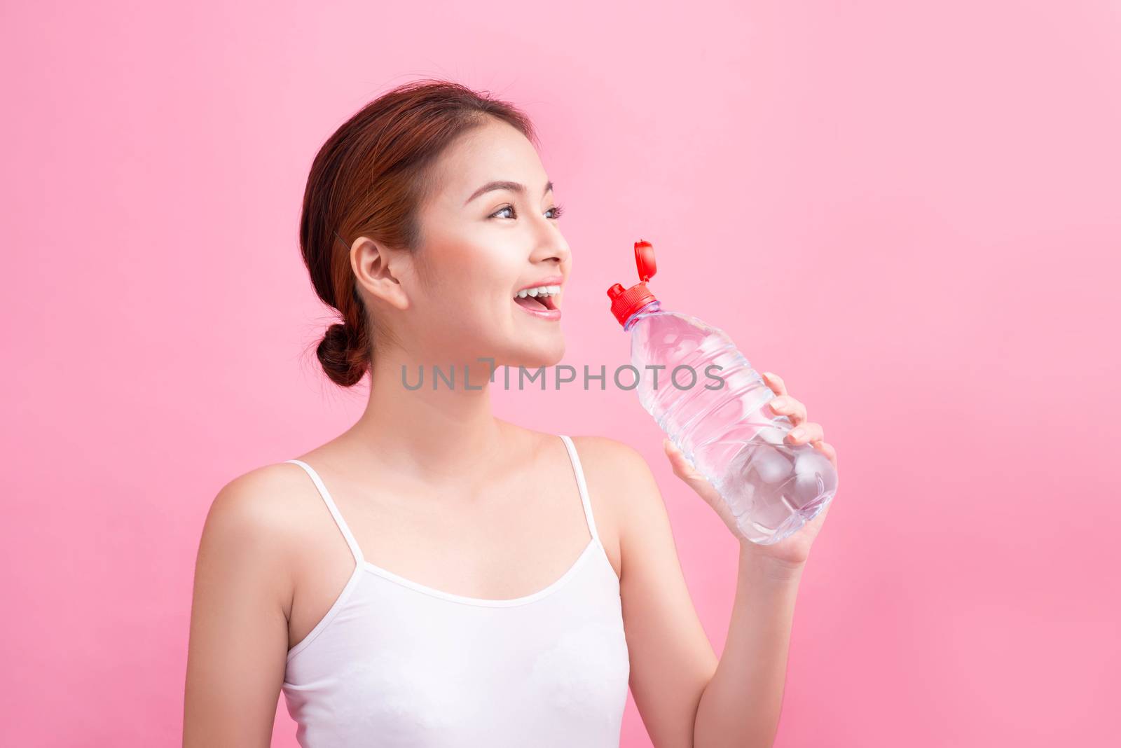 Cheerful beautiful sporty asian young woman holding a bottle of water on pink background.