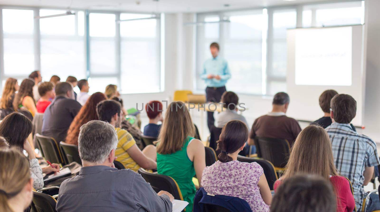 Business and entrepreneurship symposium. Speaker giving a talk at business meeting. Audience in conference hall. Rear view of unrecognized participant in audience.