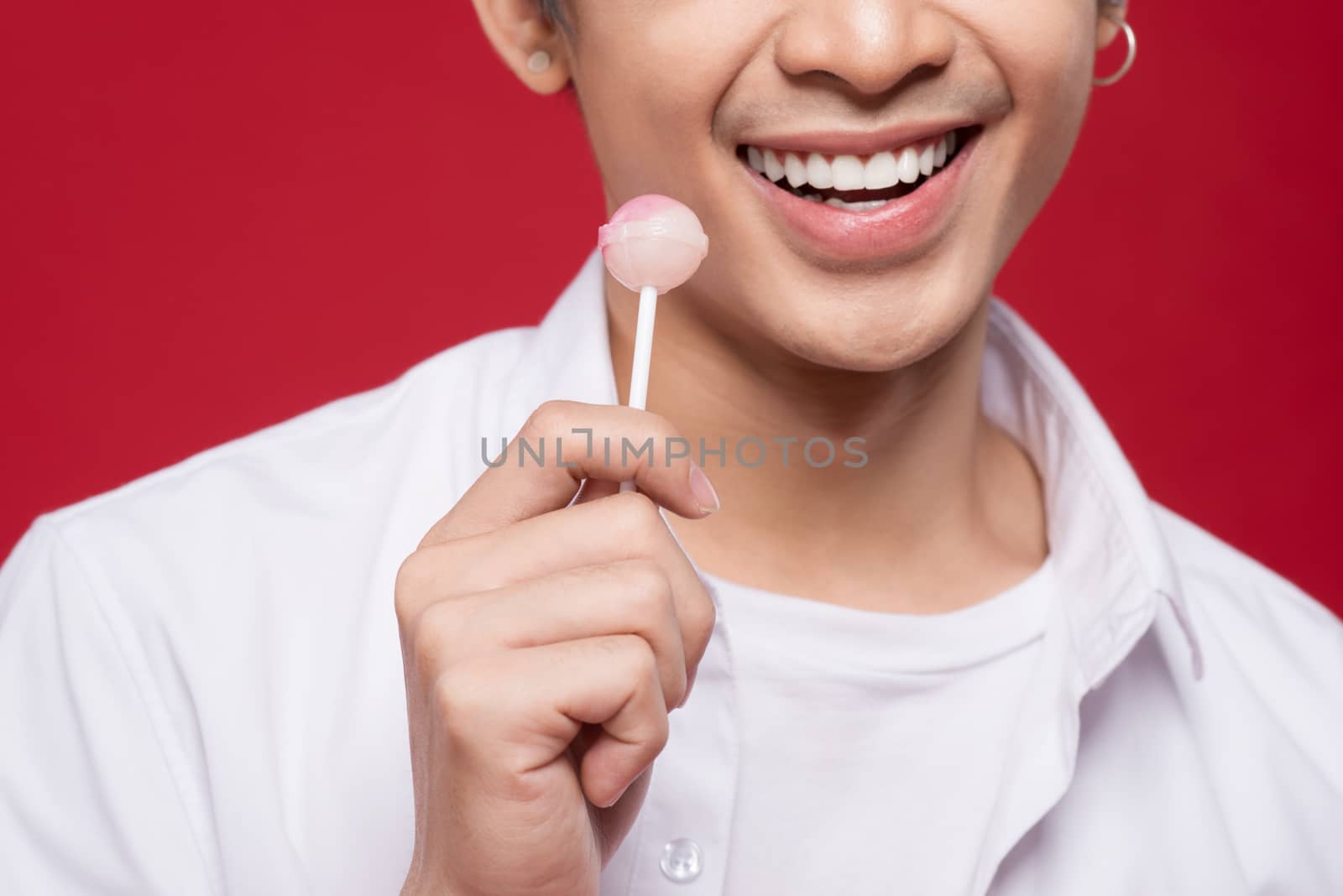 Portrait of a young stylish asian man licking a shiny lollipop over red background.
