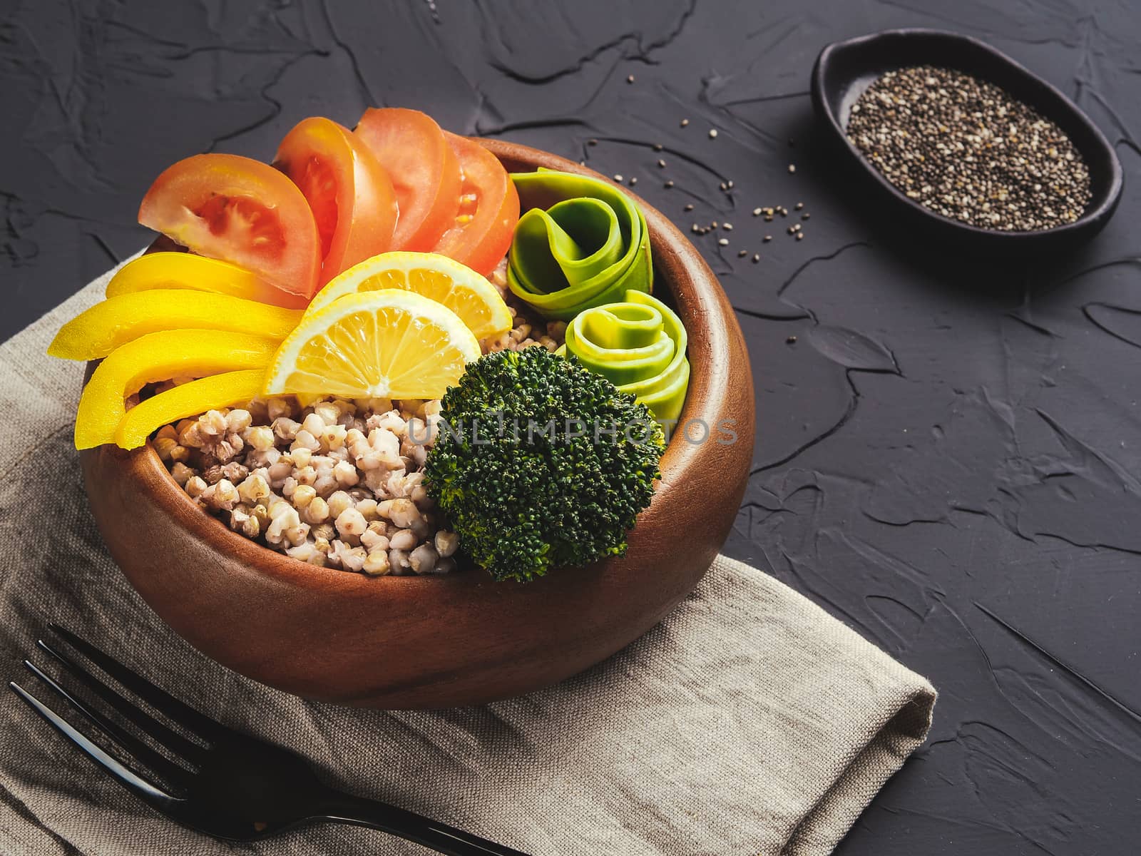 Close up view of vegetarian buddha bowl with green buckwheat, broccoli, avocado, tomatoes and yellow sweet pepper paprika on dark concrete background with copy space