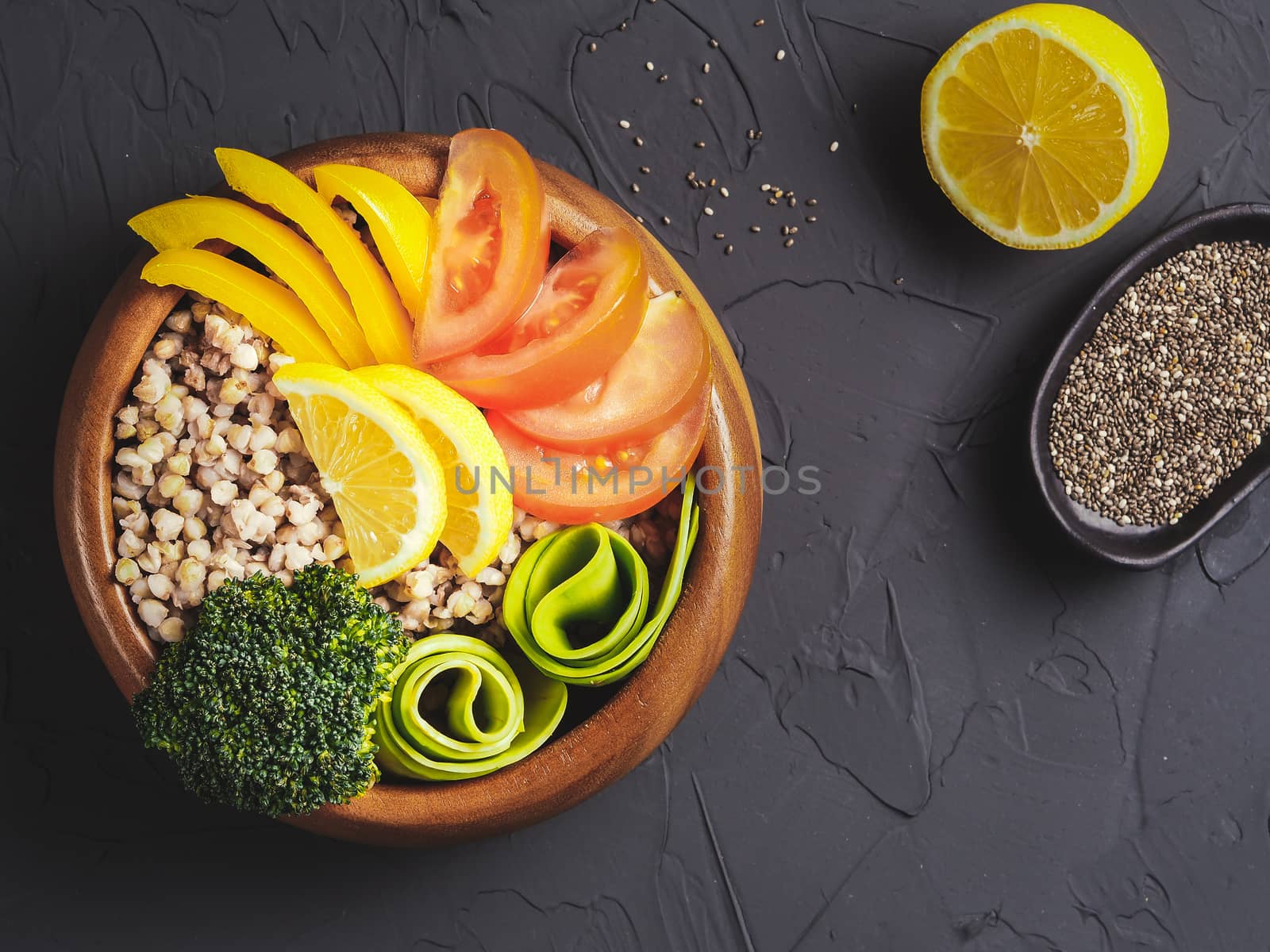 Top view of vegetarian buddha bowl with green buckwheat, broccoli, avocado, tomatoes and yellow sweet pepper paprika on dark concrete background with copy space