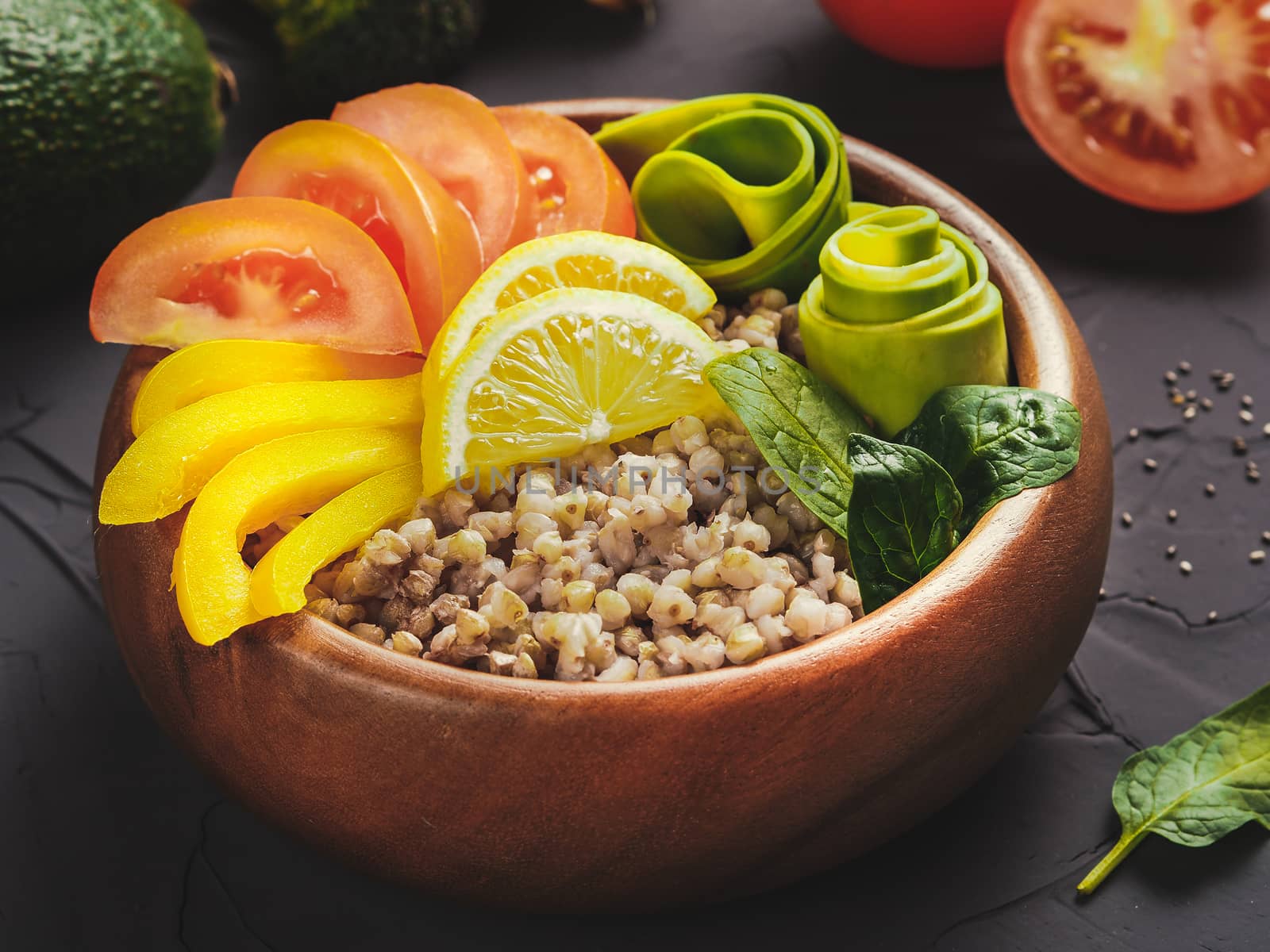 Close up view of vegetarian buddha bowl with green buckwheat, spinach, avocado, tomatoes and yellow sweet pepper paprika on dark concrete background with copy space
