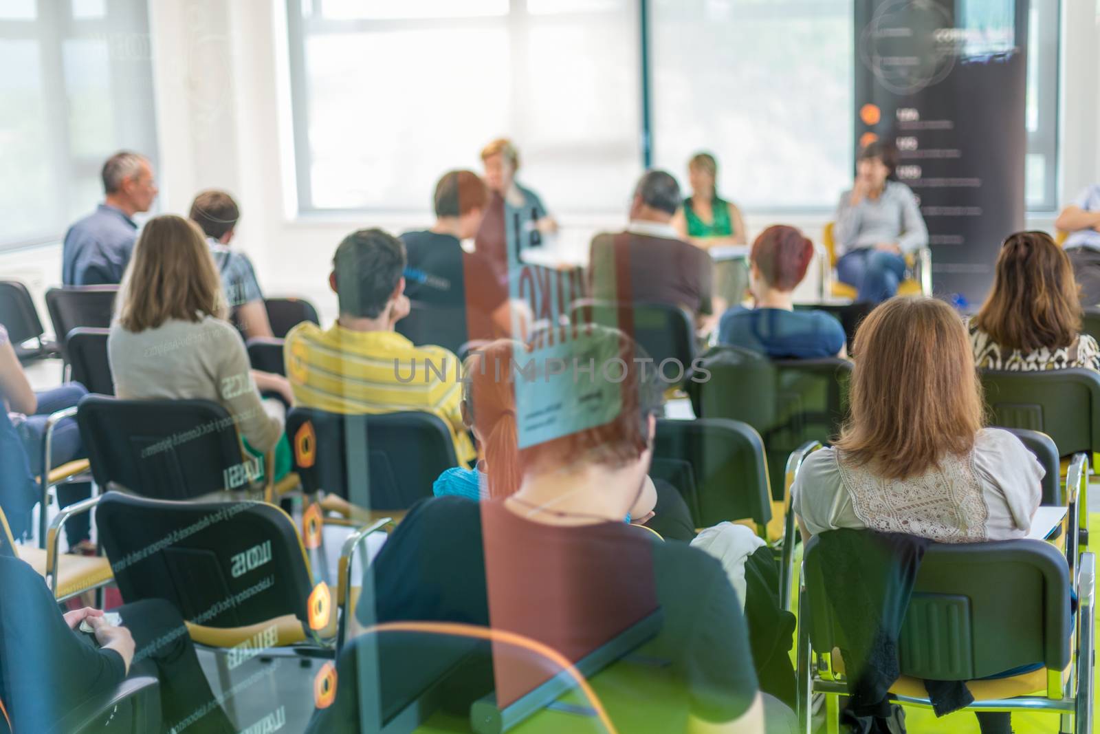 Round table discussion at business and entrepreneurship symposium. Audience in conference hall. Lens focus on unrecognized participant in rear of audience. Shot trough glass.