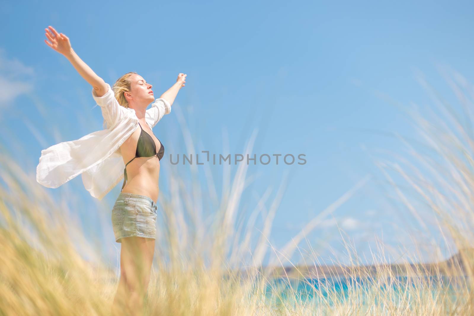 Relaxed woman, arms rised, enjoying sun, freedom and life an a beautiful beach. Young lady feeling free, relaxed and happy. Concept of vacations, freedom, happiness, enjoyment and well being.