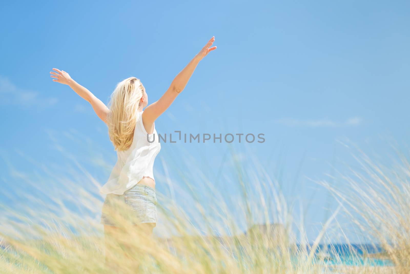 Relaxed woman, arms raised, enjoying sun, freedom and life an a beautiful beach. Young lady feeling free, relaxed and happy. Concept of vacations, freedom, happiness, enjoyment and well being.