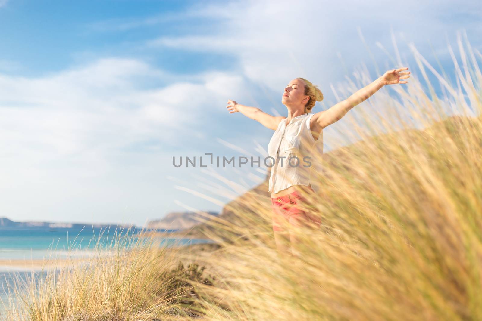 Relaxed woman, arms rised, enjoying sun, freedom and life an a beautiful beach. Young lady feeling free, relaxed and happy. Concept of vacations, freedom, happiness, enjoyment and well being.