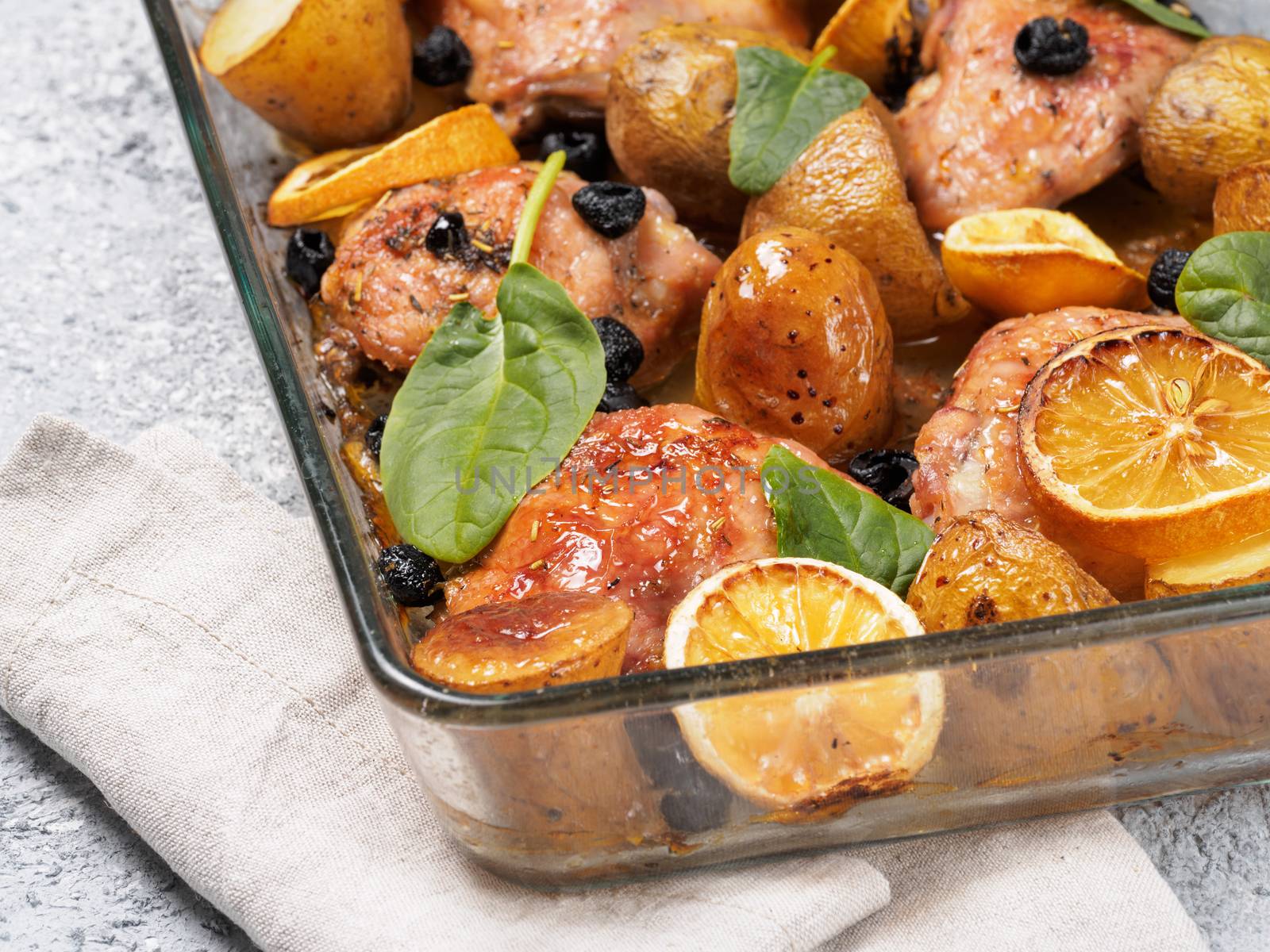 Close up view of chicken thighs with potatoes, lemon and black olives, cooked in oven on gray concrete background. Baked chicken leg quarter in heat-proof glass.