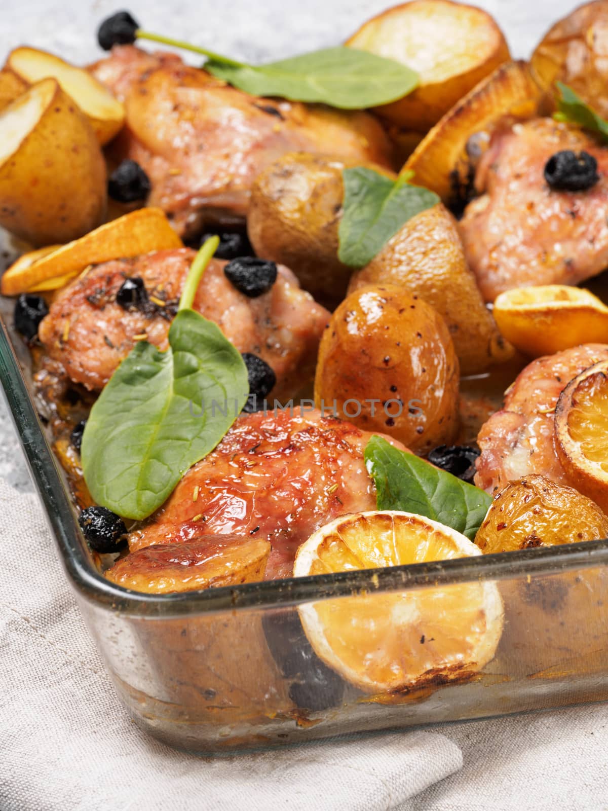 Close up view of chicken thighs with potatoes, lemon and black olives, cooked in oven on gray concrete background. Baked chicken leg quarter in heat-proof glass.