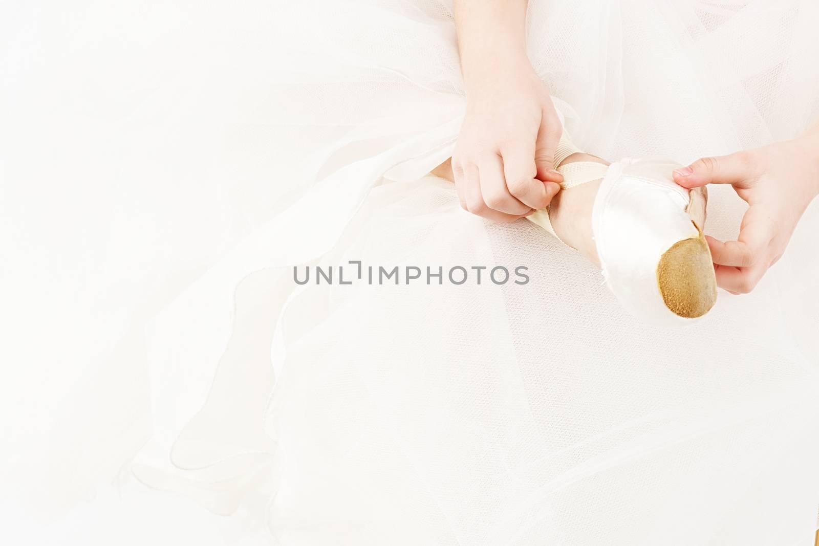 A small ballerina dresses or takes off her pointe isolated on white background. Slender little ballerina girl in white dress and in pointe shoes