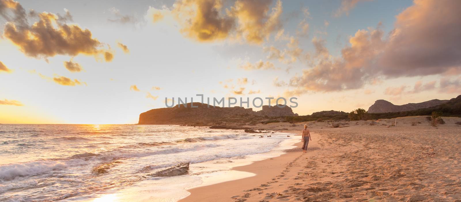 Woman walking on sandy beach at golden hour. Seashore sunset walk, Falasarna, Crete, Greece.