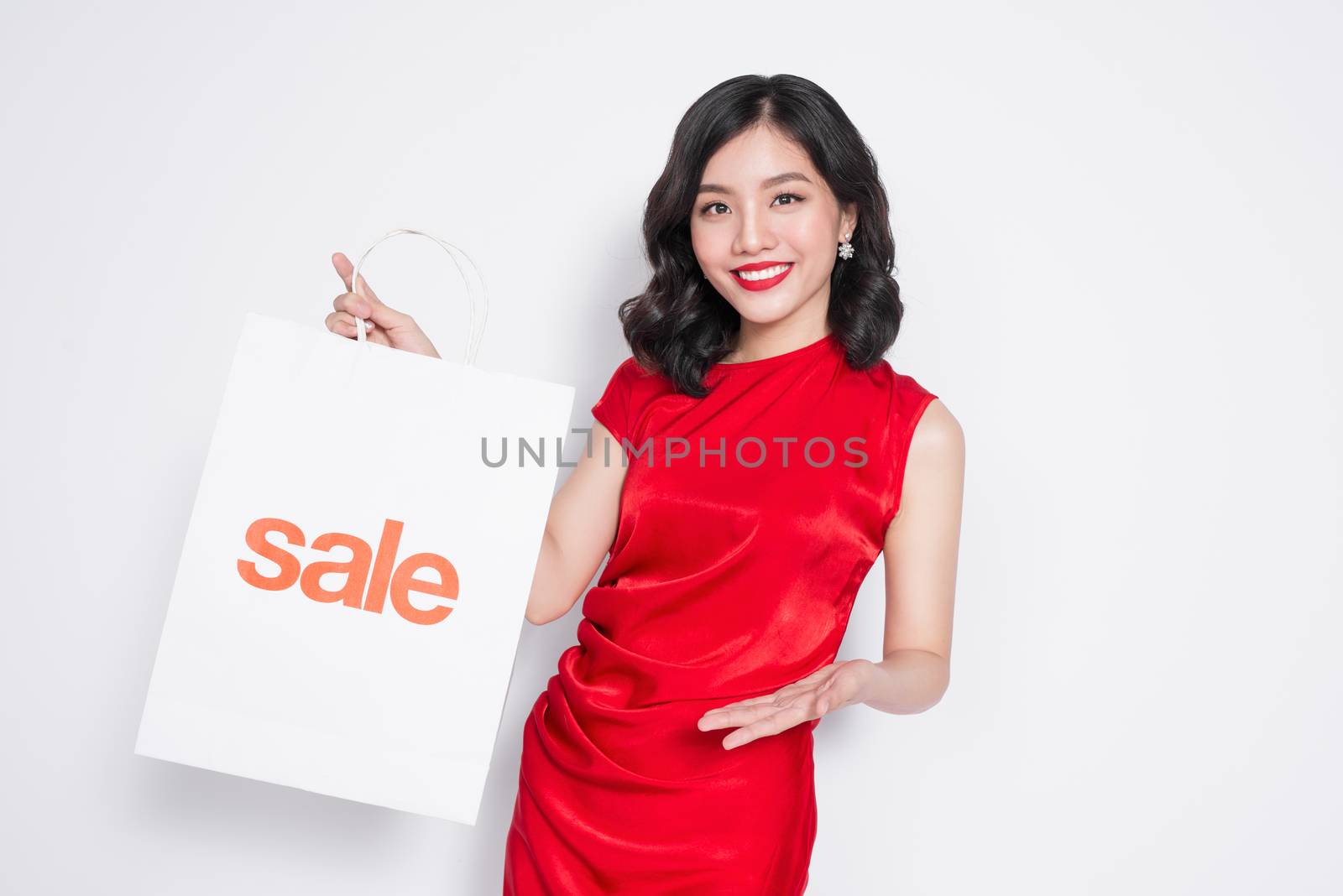 Beautiful asian woman wearing a red dress with shopping bag standing over white.