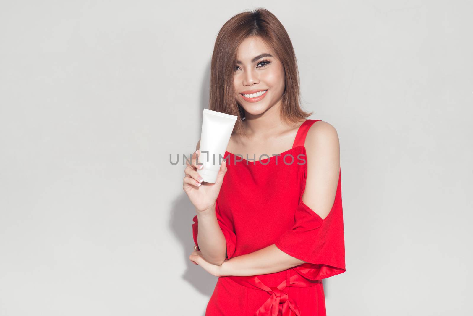 Portrait of cheerful smiling fashion girl in red dress holding cosmetic plastic tube
