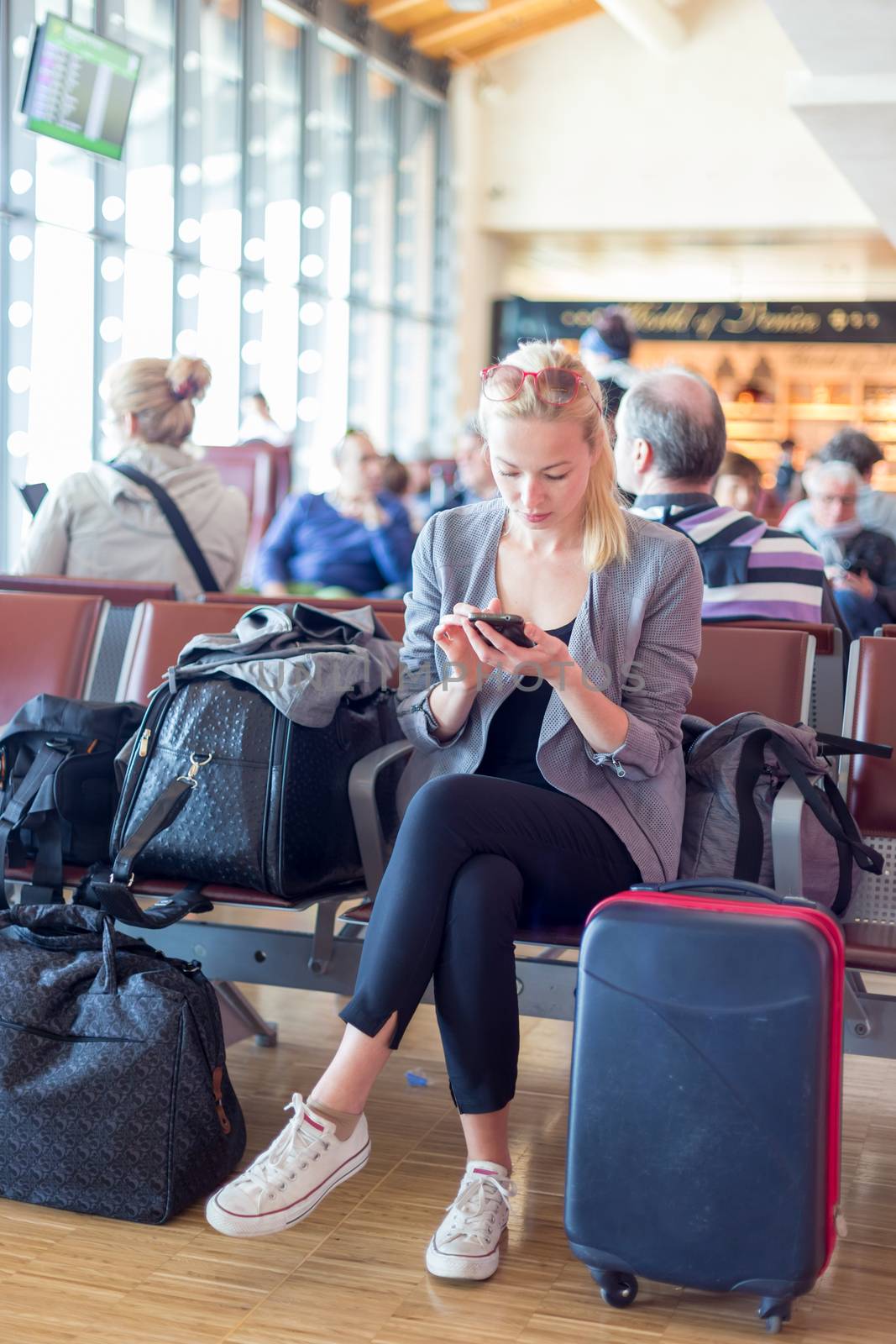 Casual blond young woman using her cell phone while waiting to board a plane at the departure gates. Wireless network hotspot enabling people to access internet conection. Public transport.