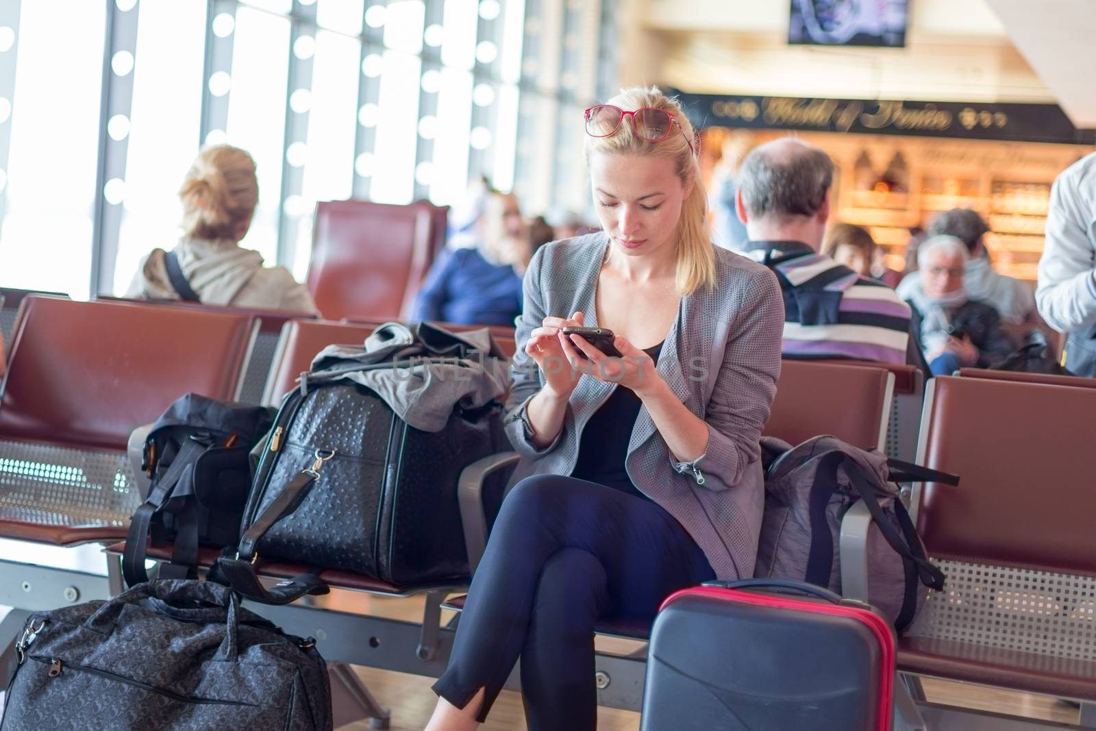 Casual blond young woman using her cell phone while waiting to board a plane at the departure gates. Wireless network hotspot enabling people to access internet conection. Public transport.