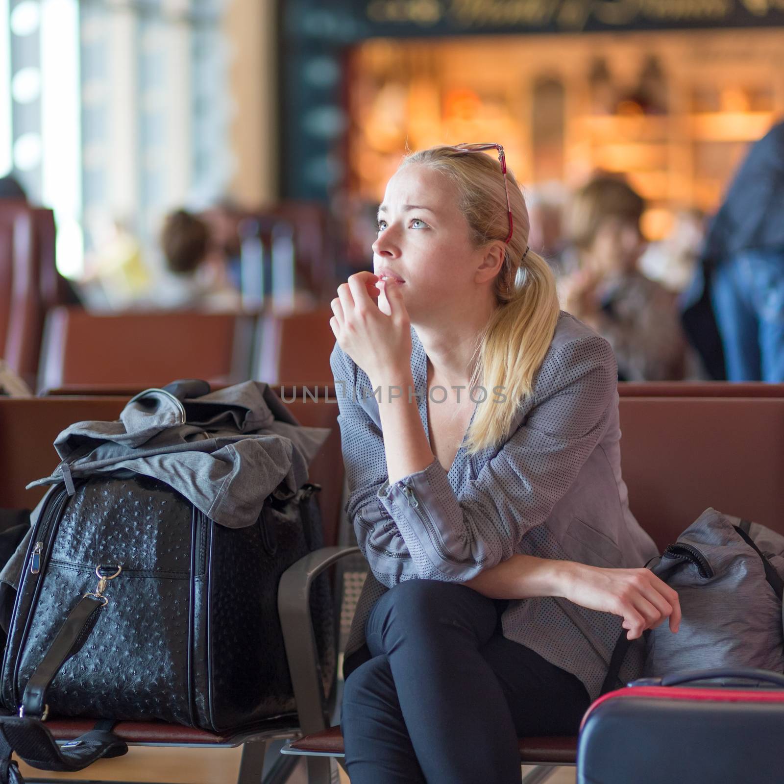 Young blond caucsian woman waiting on airport terminal full of passenegers for her flight to depart.