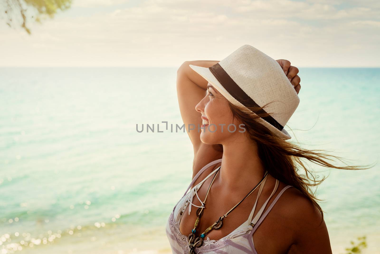 Beautiful young woman with summer hat enjoying on the beach. She is smiling and pensive looking away. 