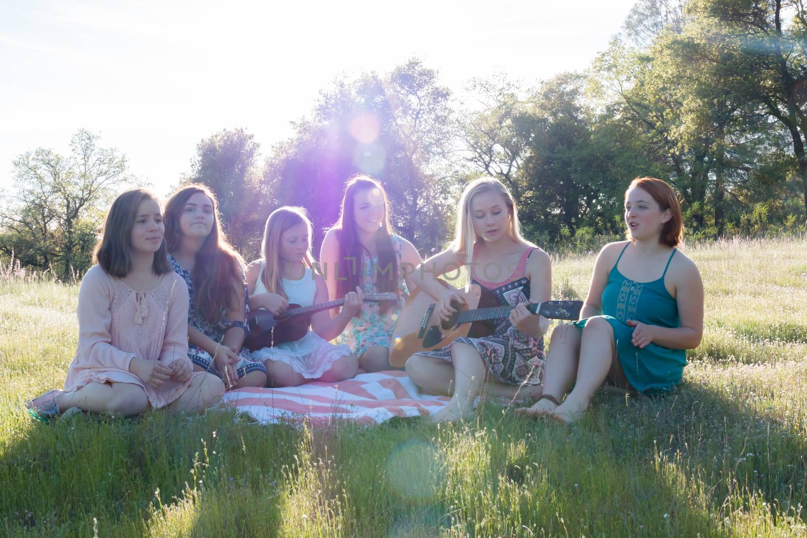 Young Girls Sitting Together in Grassy Field Singing and Playing Musical Instruments With Sunlight Overhead