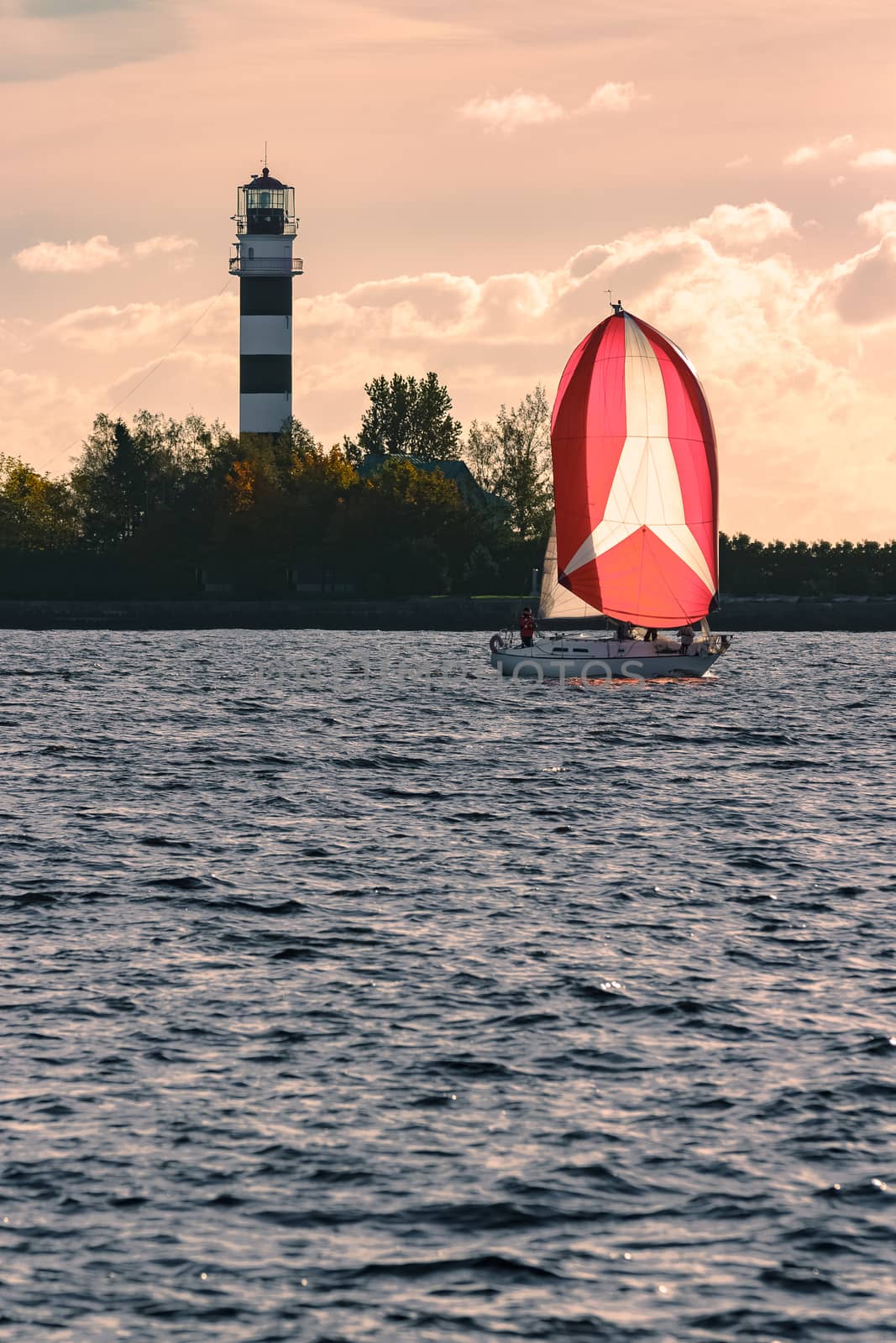 Red sailboat sailing past the big lighthouse in evening, Latvia