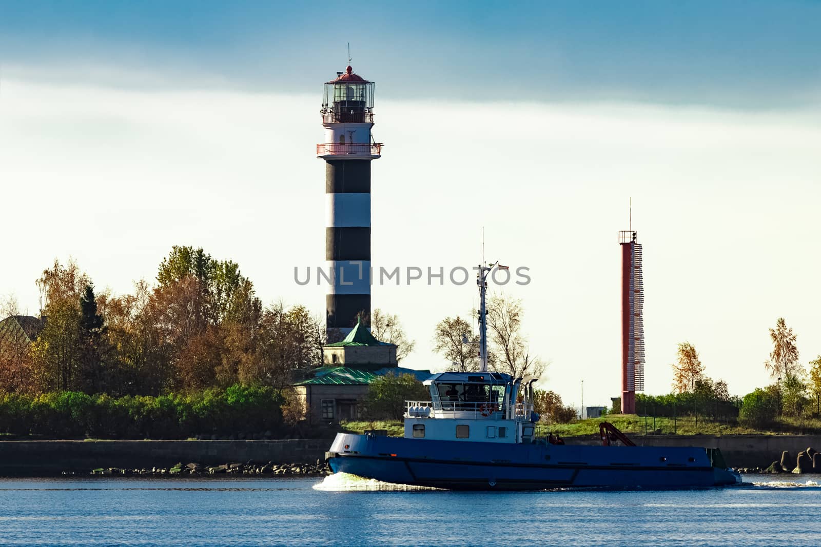 Blue small tug ship sailing past the big lighthouse at sunny day