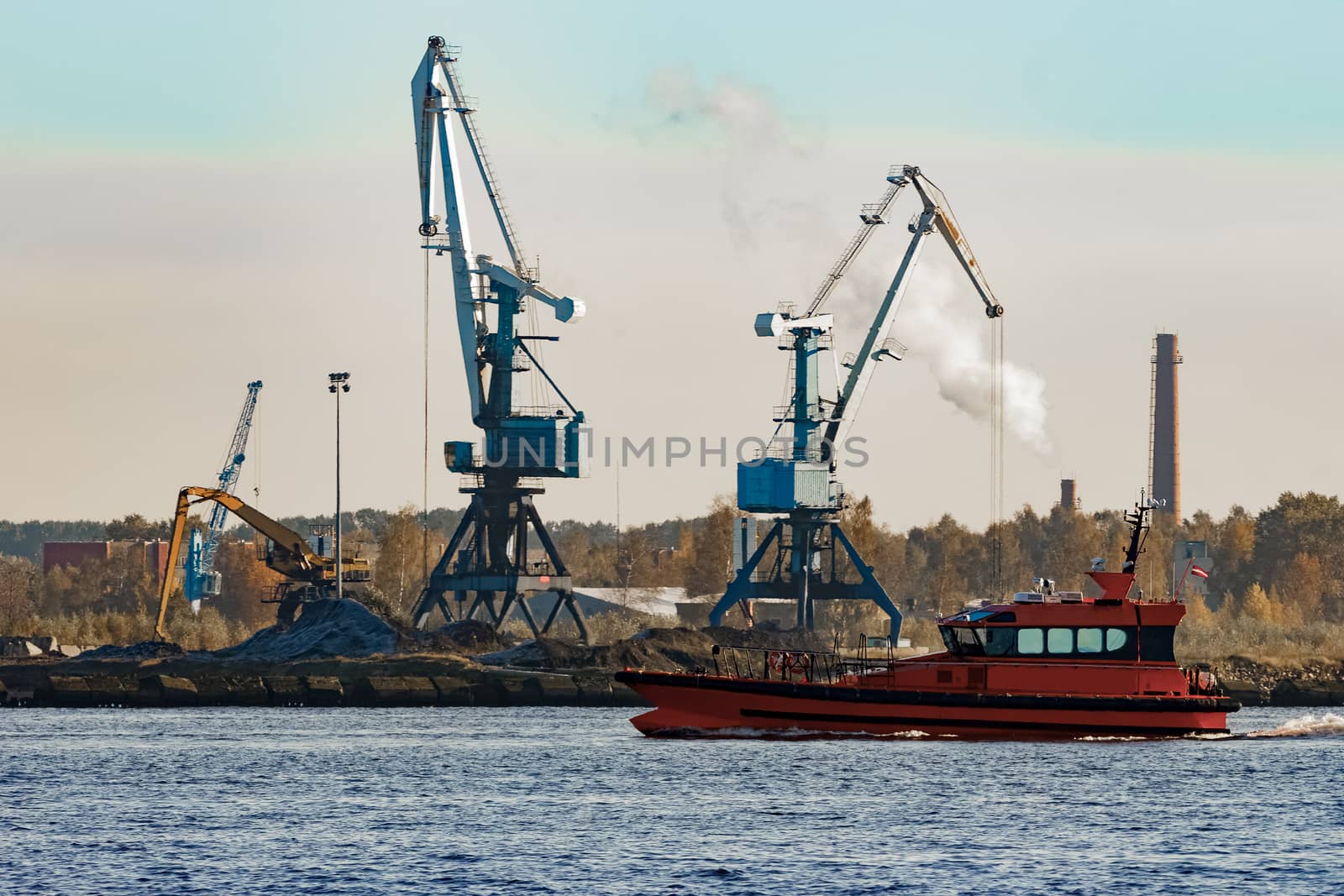 Orange pilot ship sailing against the cargo cranes