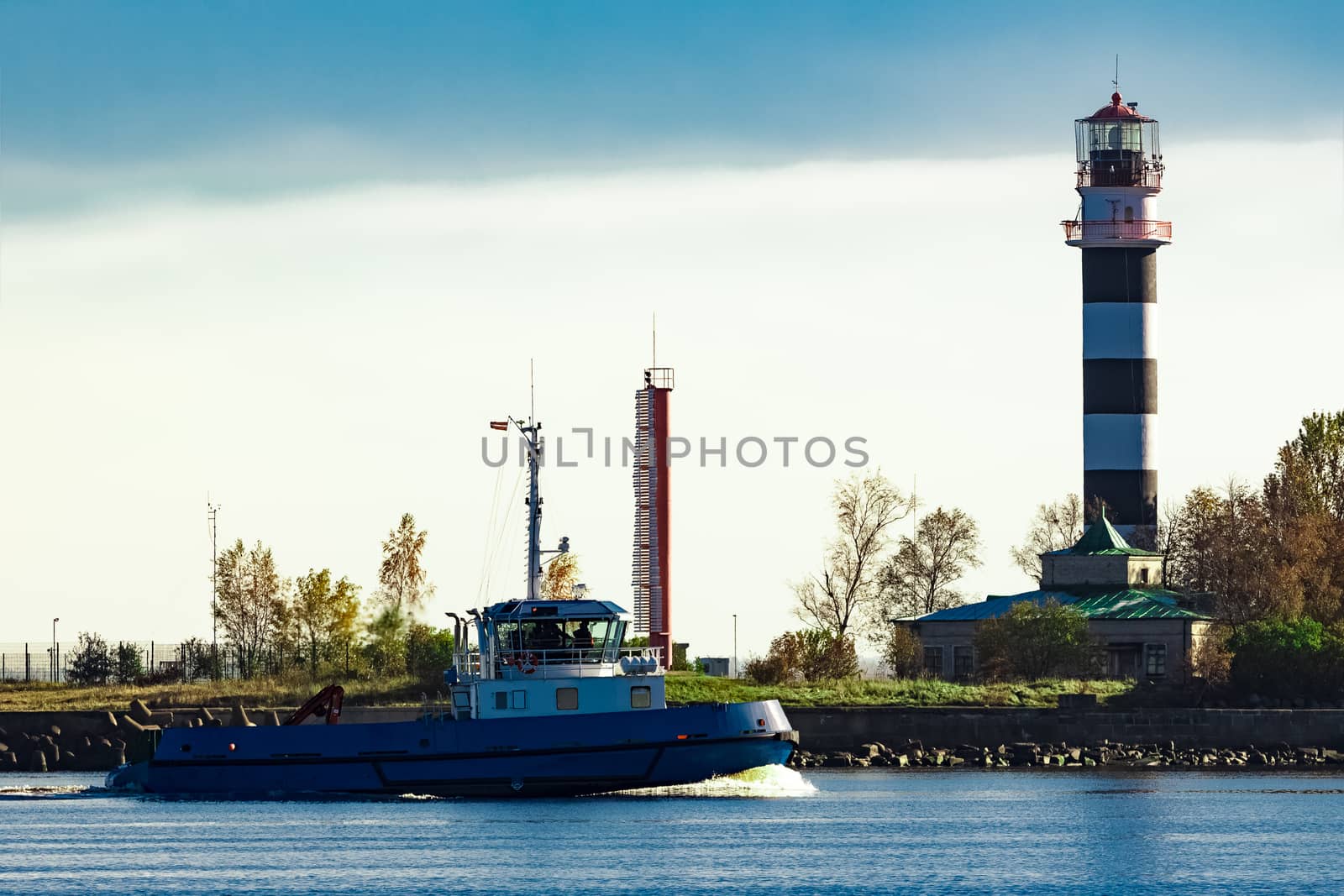 Blue small tug ship sailing past the big lighthouse at sunny day