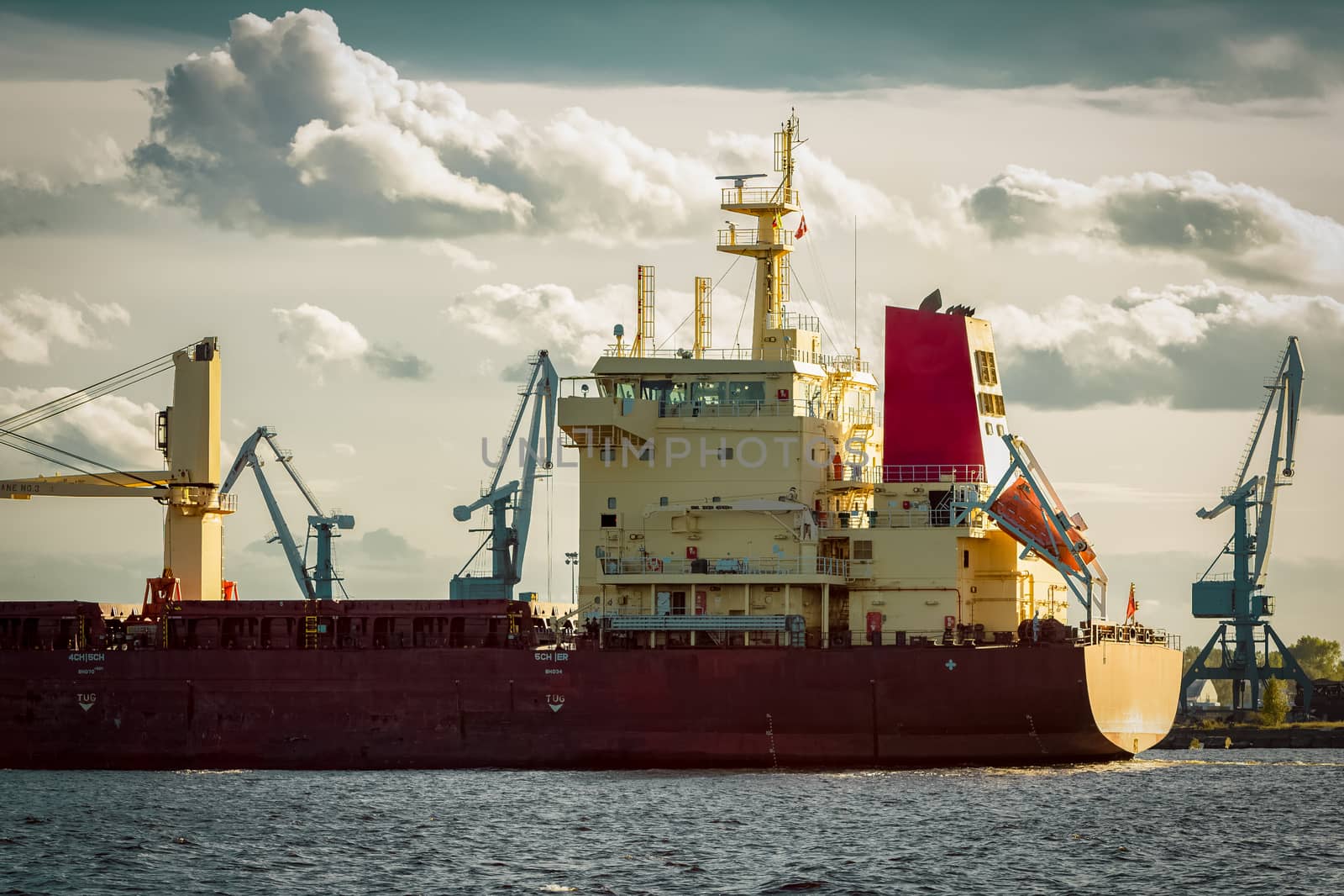 Red cargo ship's stern against port cranes