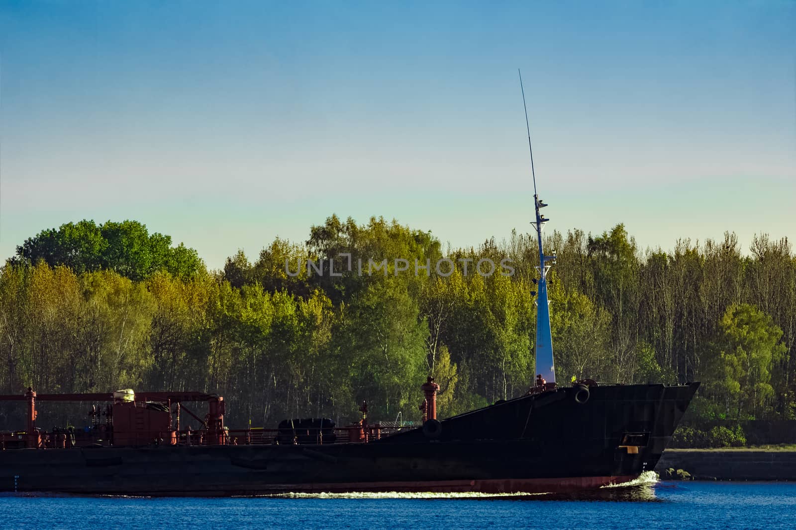 Black cargo oil tanker's bow against summer green trees