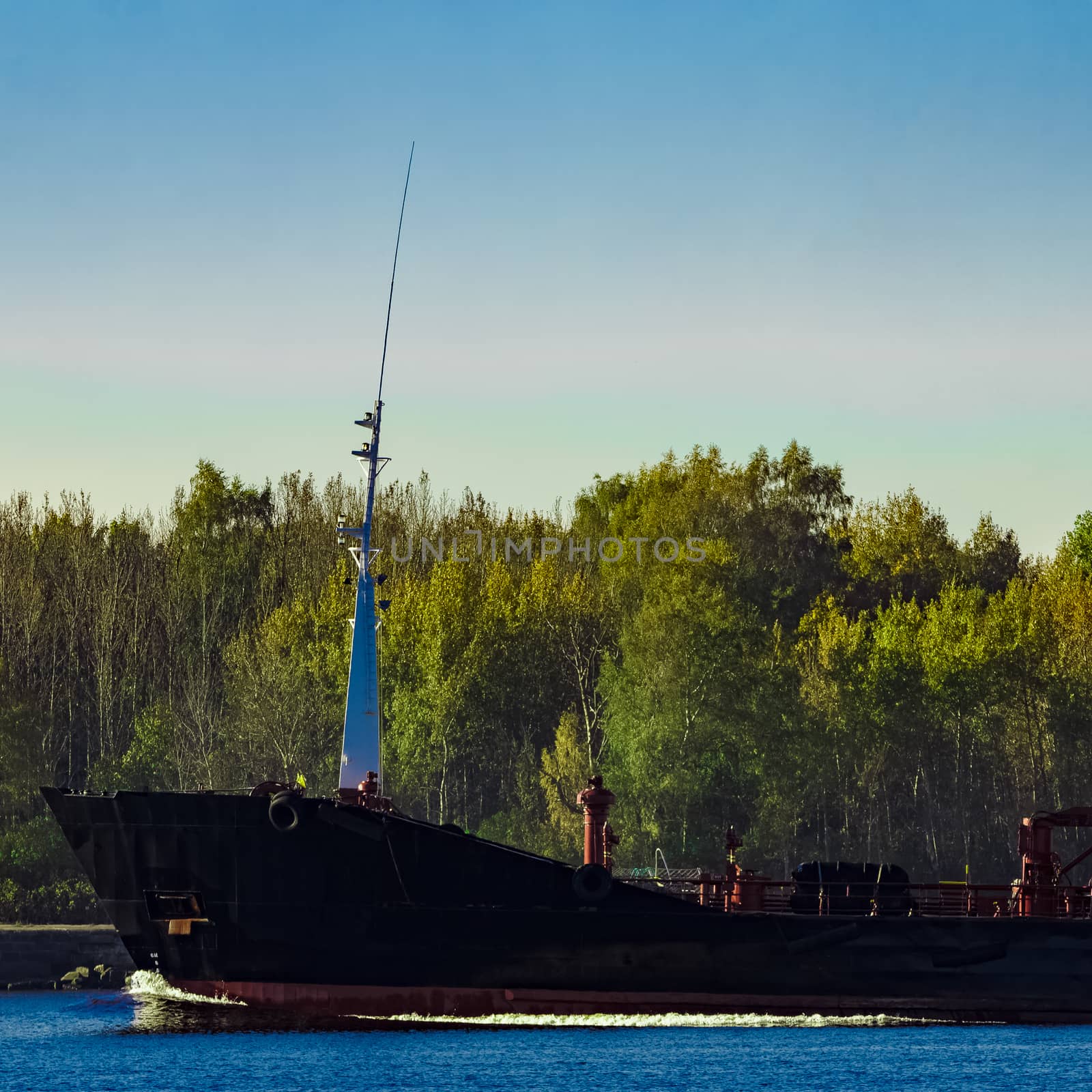 Black cargo oil tanker's bow against summer green trees