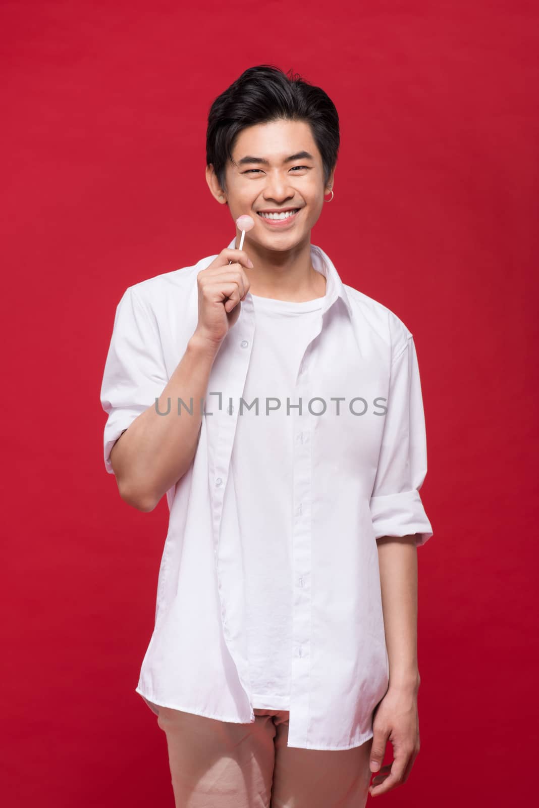 Portrait of a young stylish asian man licking a shiny lollipop over red background.