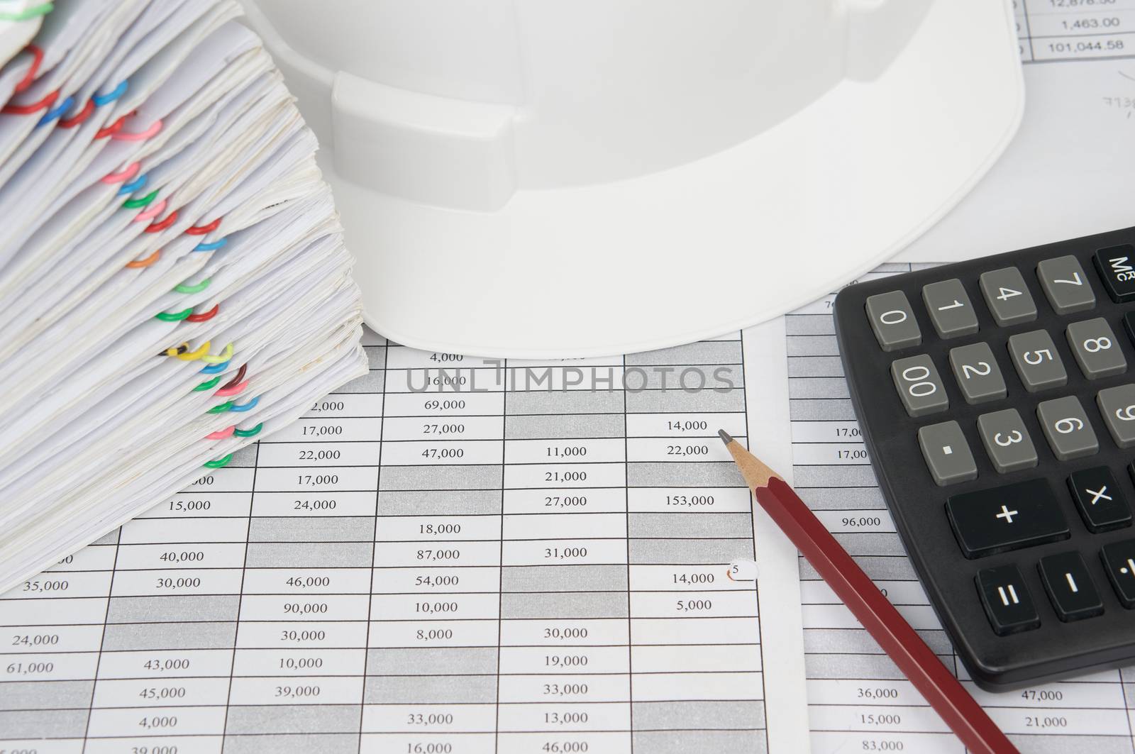 Pencil and calculator on finance account have blur white engineer hat and pile overload document of report and receipt with colorful paperclip as background.