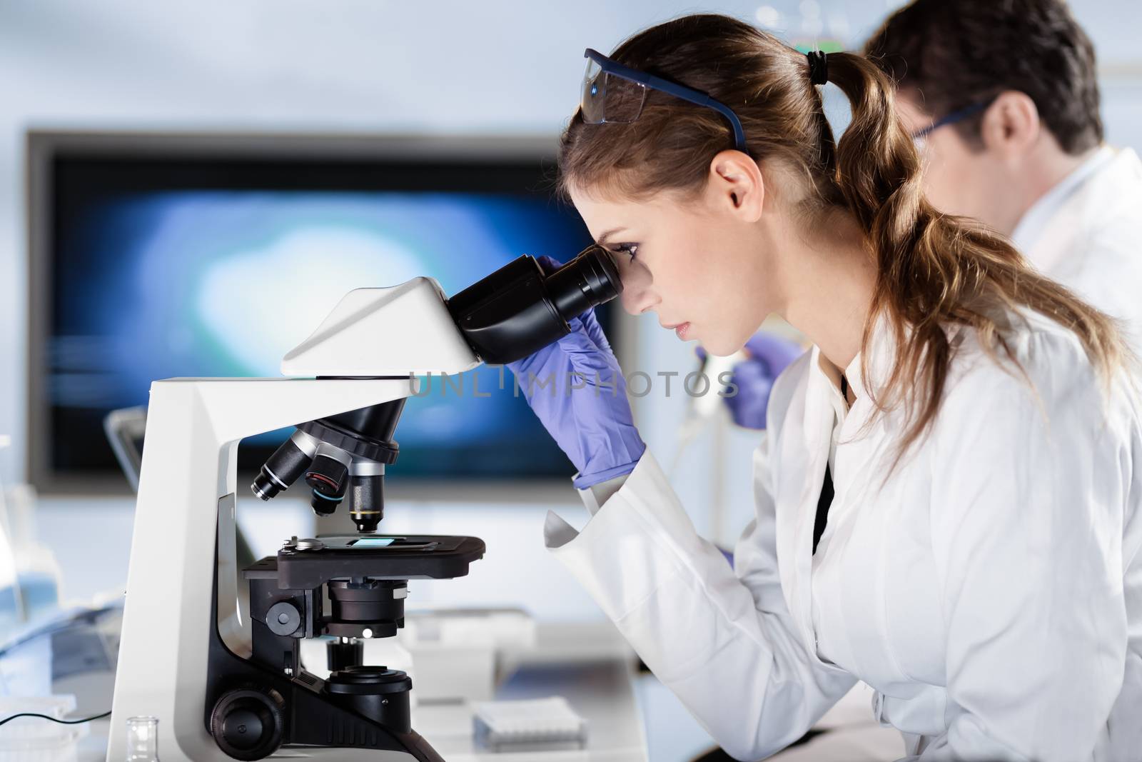 Life scientists researching in laboratory. Attractive female young scientist and her post doctoral supervisor microscoping in their working environment. Healthcare and biotechnology.