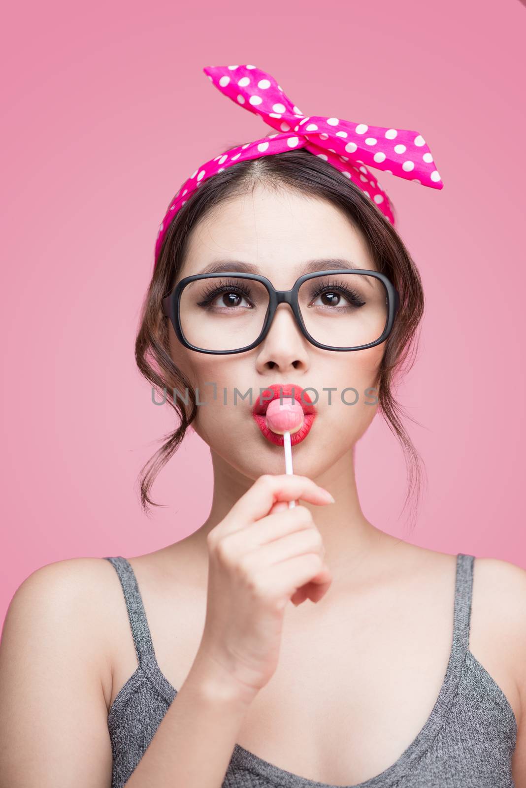 Portrait of beautiful asian woman eating heart shape lollipop, dressed and makeup in pin-up style on pink background.