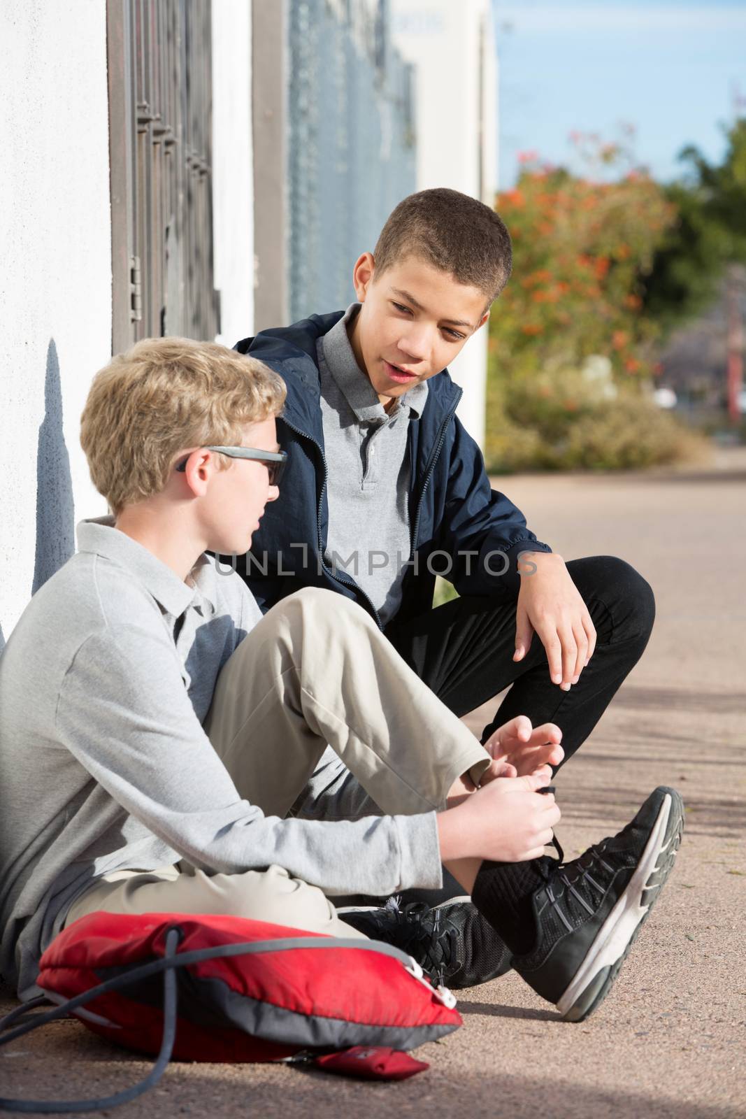 Two male teenage students hanging out at side of wall talking. Backpack on ground.