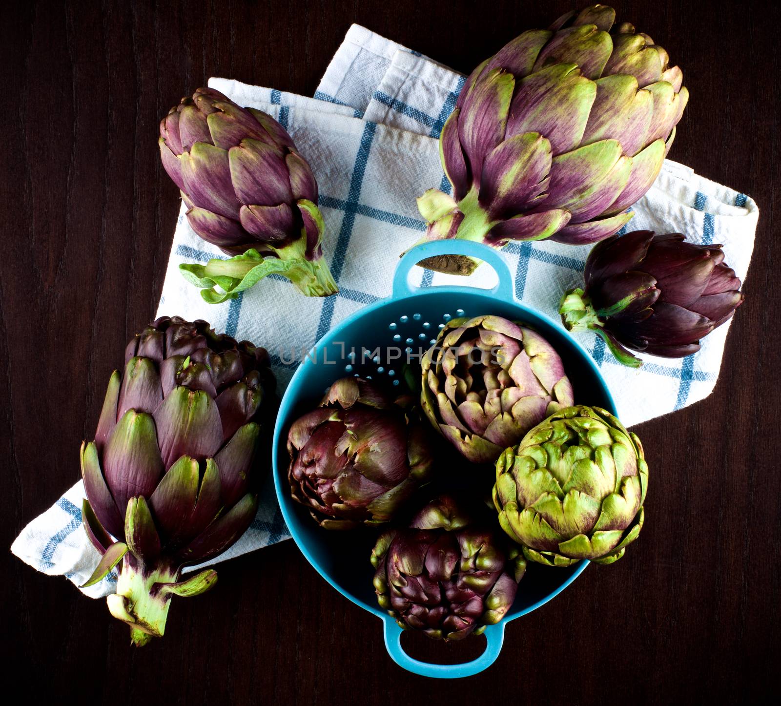 Stack of Perfect Raw Artichokes in Blue Colander closeup on Checkered Napkin closeup on Dark Wooden background. Top View