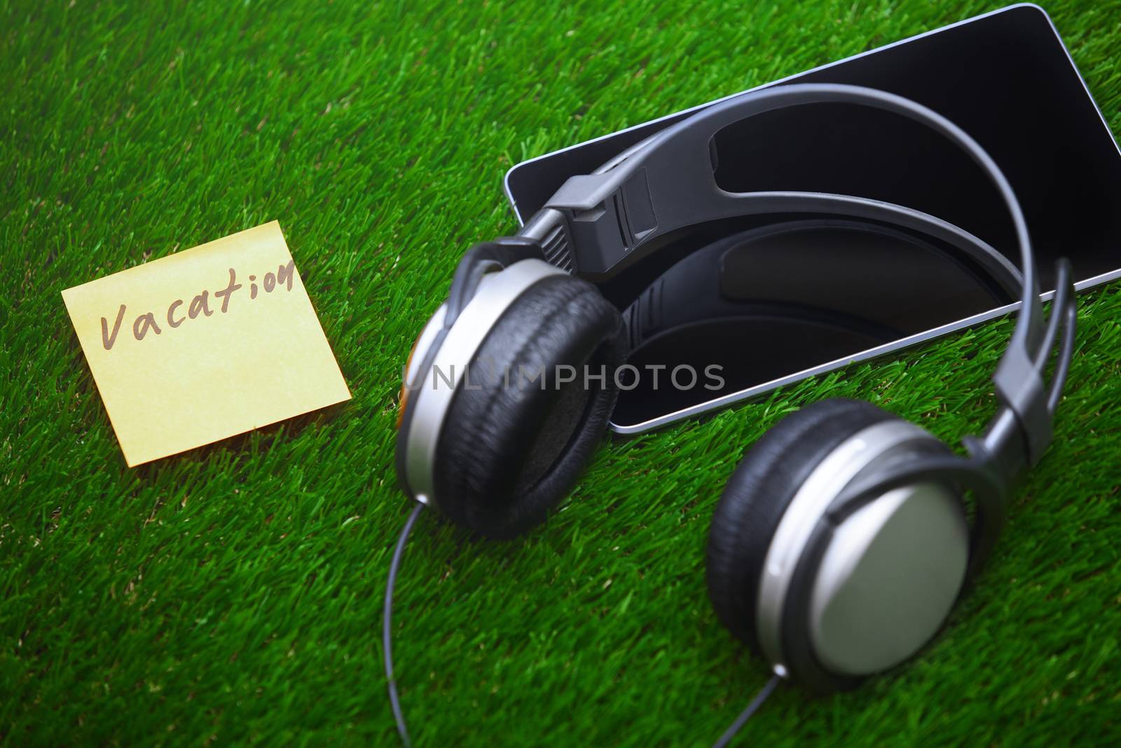 Vacation text on sticky note on a grass with headphones and digital tablet