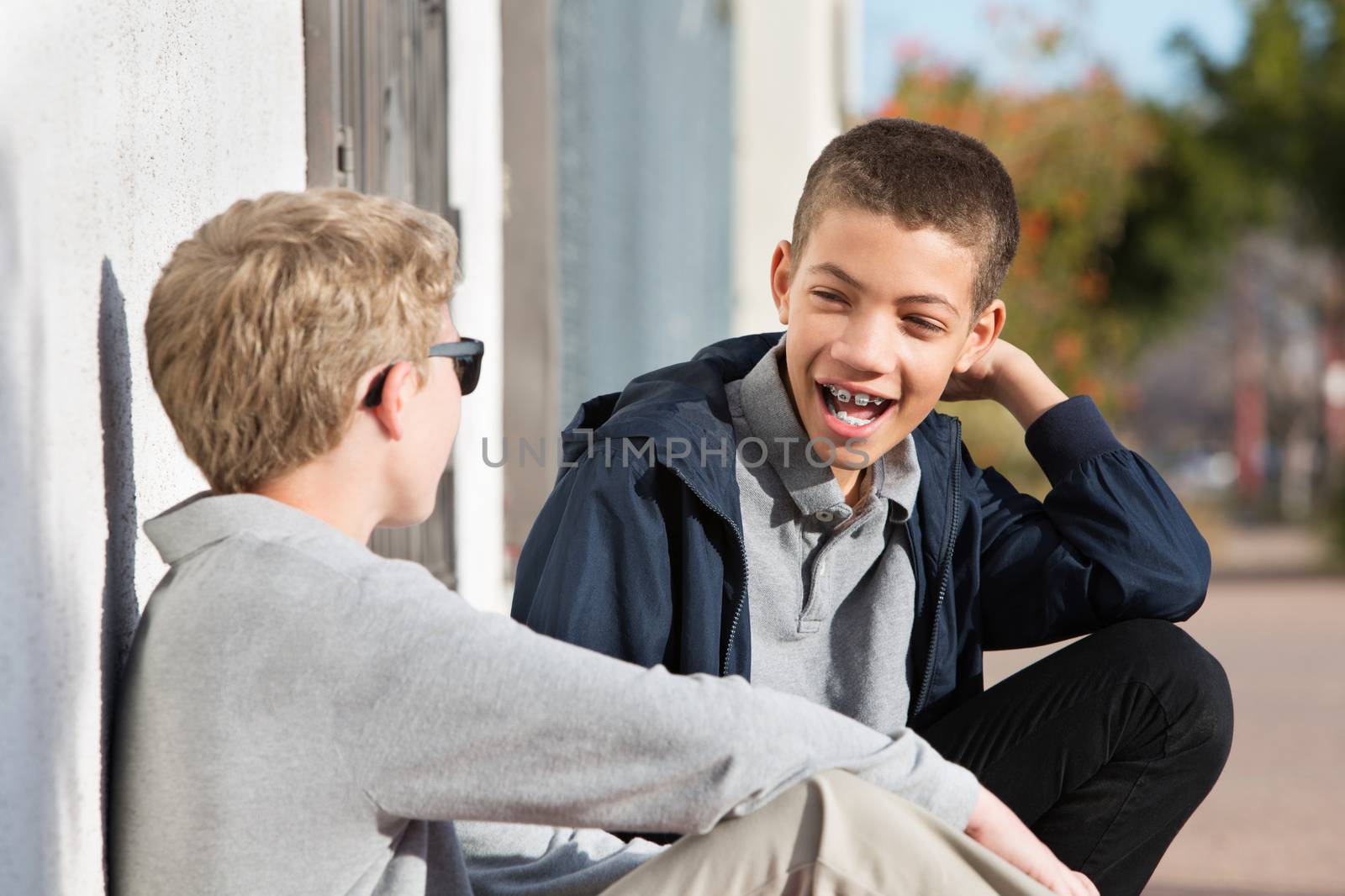 Laughing male teenager with braces beside friend sitting against wall outside