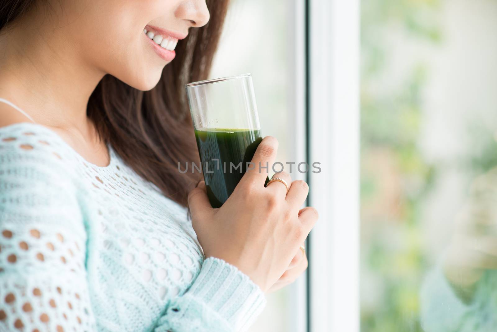 Smiling young asian woman drinking green fresh vegetable juice or smoothie from glass at home
