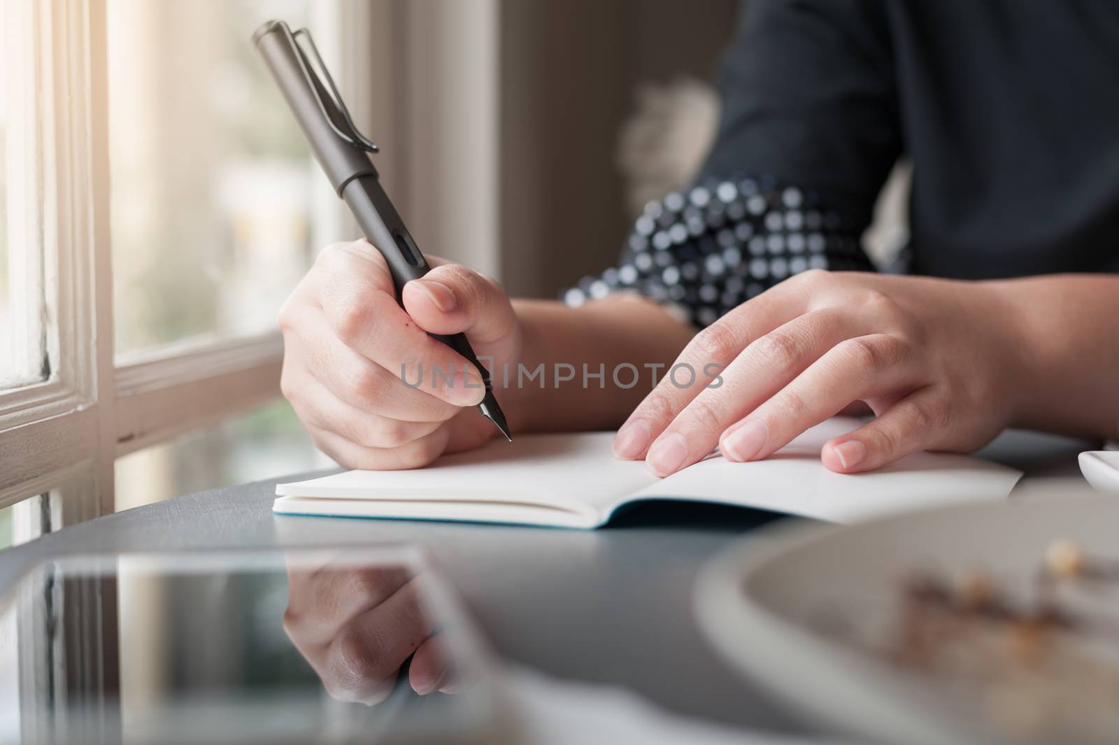 Woman hand holding pen while writing on small notebook beside window. Freelance journalist working at home concept.