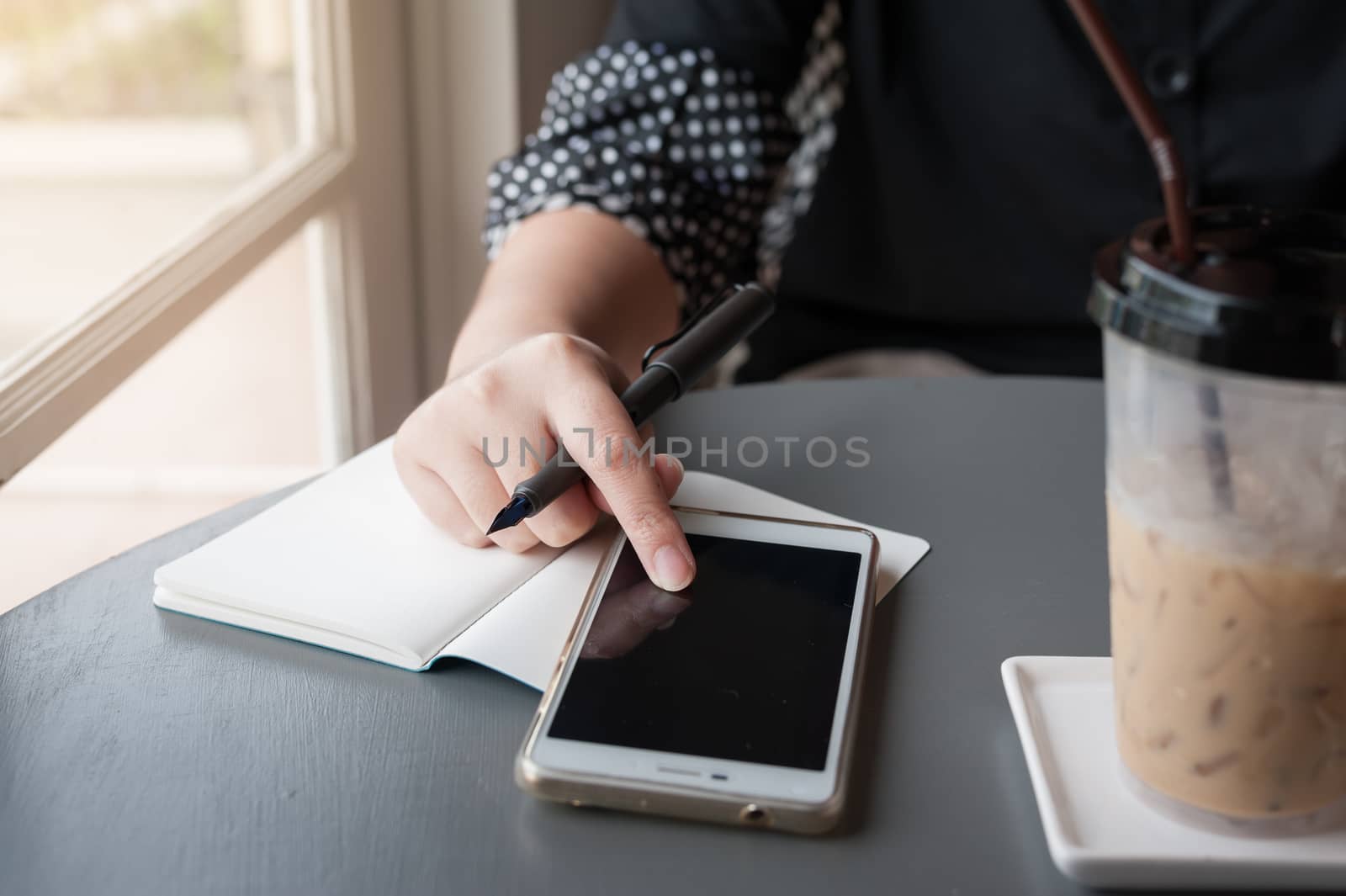 Woman hand touching on smartphone screen while writing something in coffee shop. Working from anywhere concept