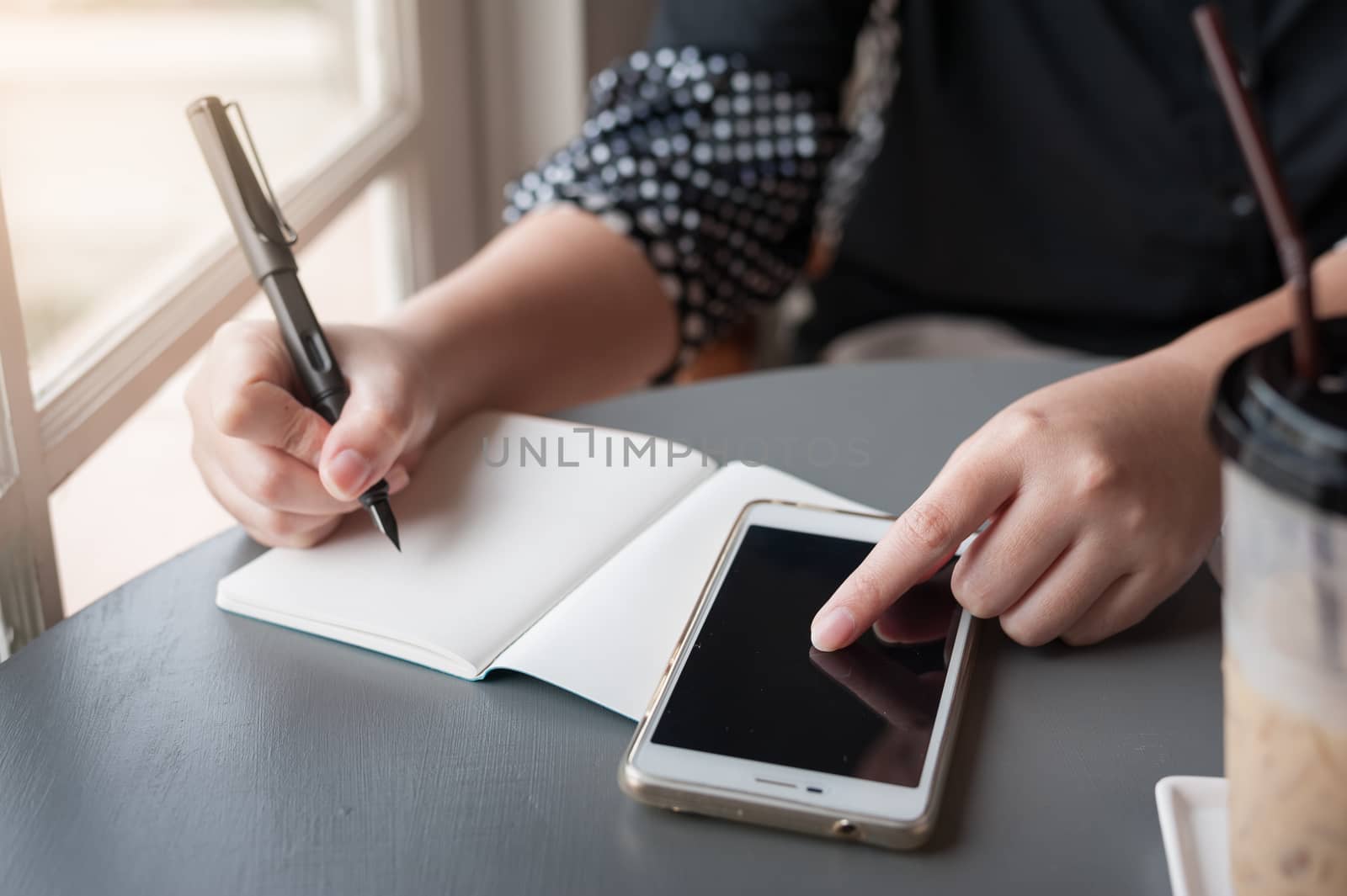 Woman hand writing a note on notebook while another hand touching on smartphone screen on table beside window. Working from anywhere concept